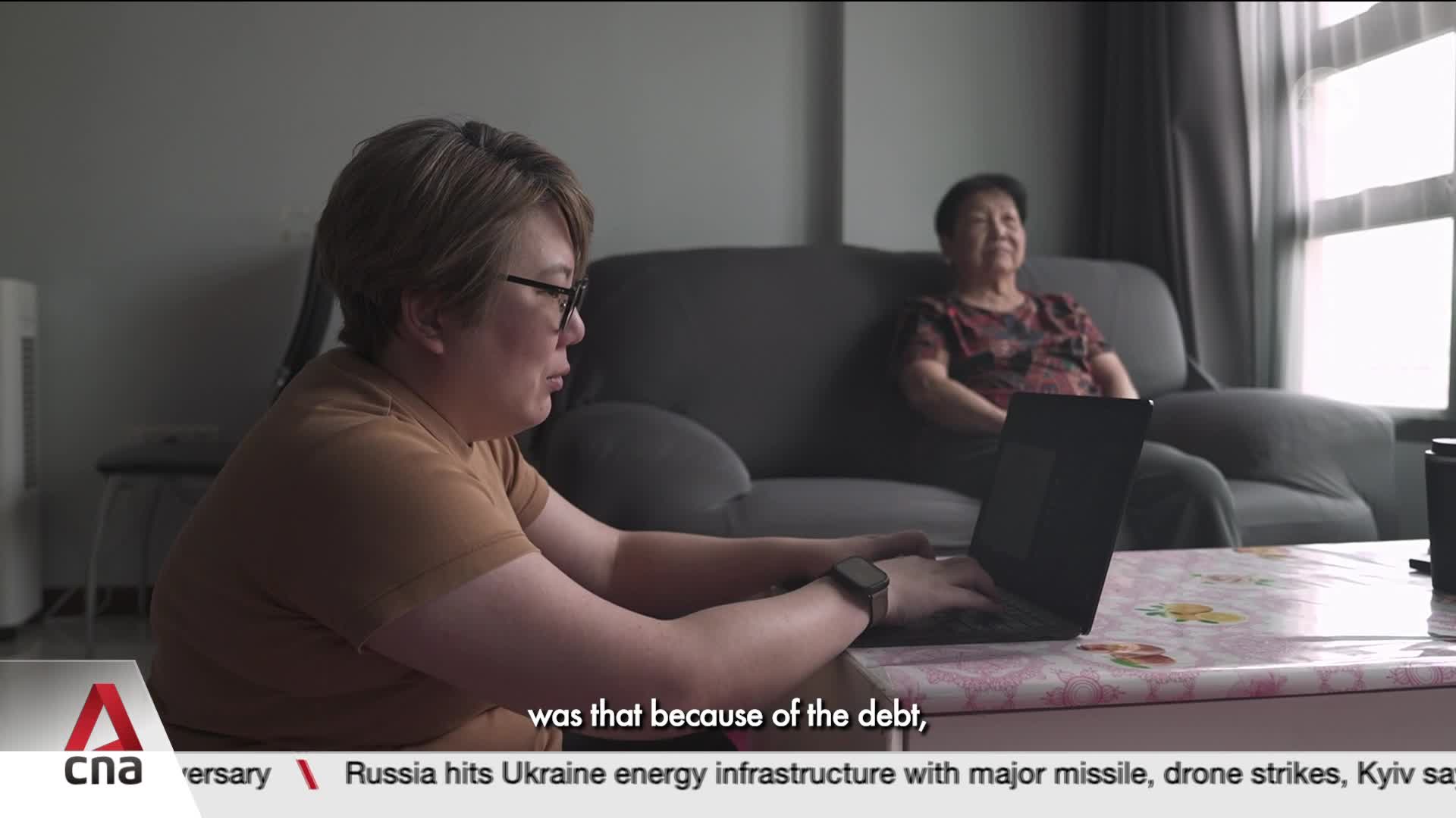 A woman in Singapore is typing on a laptop, her gaze fixed on the screen. Behind her, an older woman sits on a sofa, her expression serene. A woman in Singapore is typing on a laptop, her gaze fixed on the screen. Behind her, an older woman sits on a sofa, her expression serene.