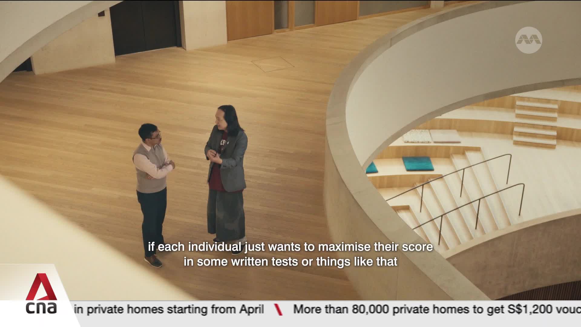 Two men stand on a polished wooden floor, engaged in conversation. One man, wearing glasses and a vest over a collared shirt, gestures with his hands. The other, with long dark hair, wears a blazer and a long skirt. Two men stand on a polished wooden floor, engaged in conversation. One man, wearing glasses and a vest over a collared shirt, gestures with his hands. The other, with long dark hair, wears a blazer and a long skirt.