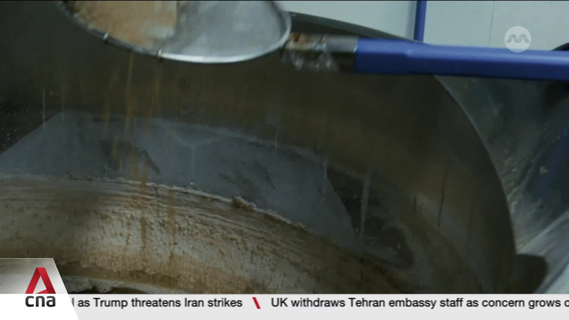 A metal scoop is tipping, letting a stream of fine, light brown powder fall into a large, industrial mixing drum. The powder lands on a bed of similar material already inside the drum.