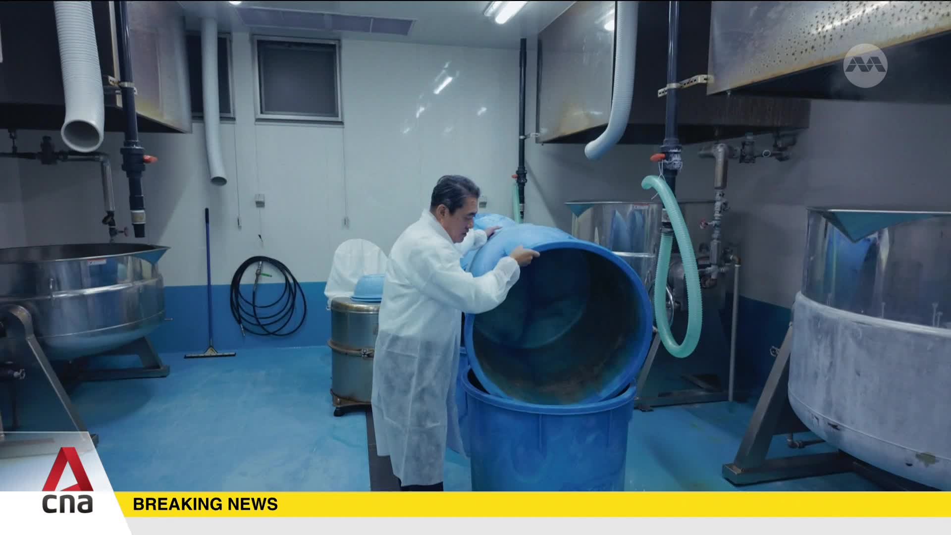 A man in a white lab coat is carefully tilting a large blue plastic lid over a matching blue bin. Industrial-sized stainless steel vats surround him in this Singapore facility.