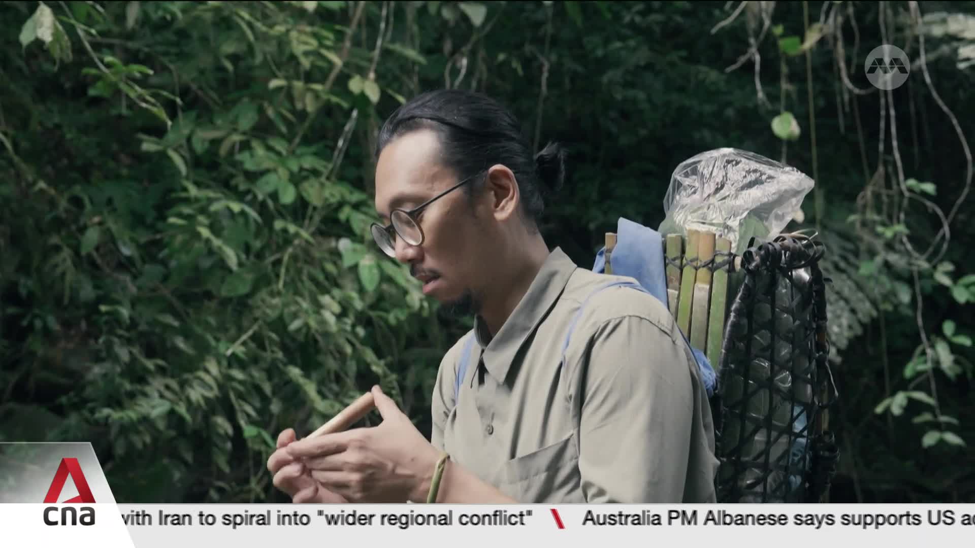 A man with glasses and his hair pulled back in a bun pauses, holding a thin wooden object. He wears a khaki shirt and a backpack constructed from bamboo and plastic.