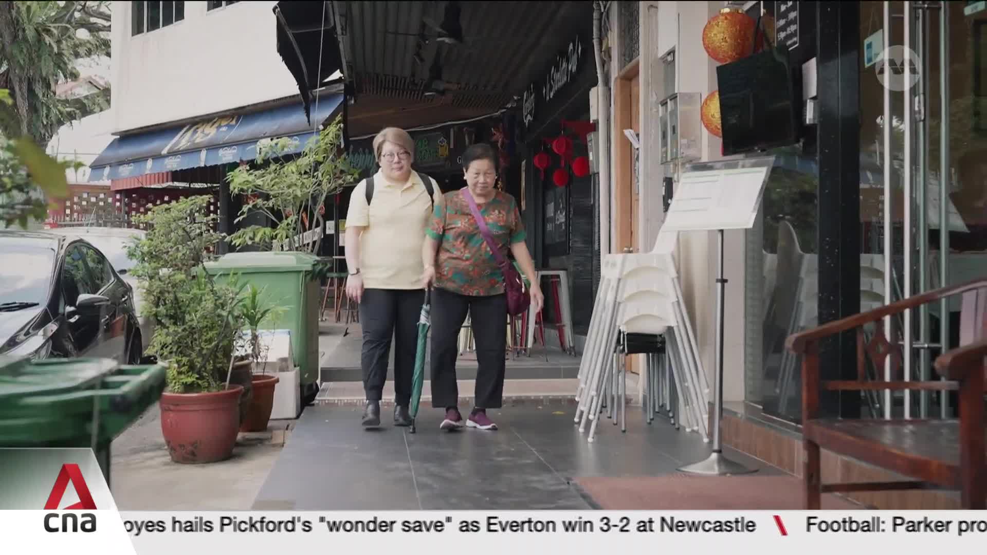 Two women walk down a Singapore sidewalk, one carrying an umbrella. They pass by shops with chairs stacked outside.