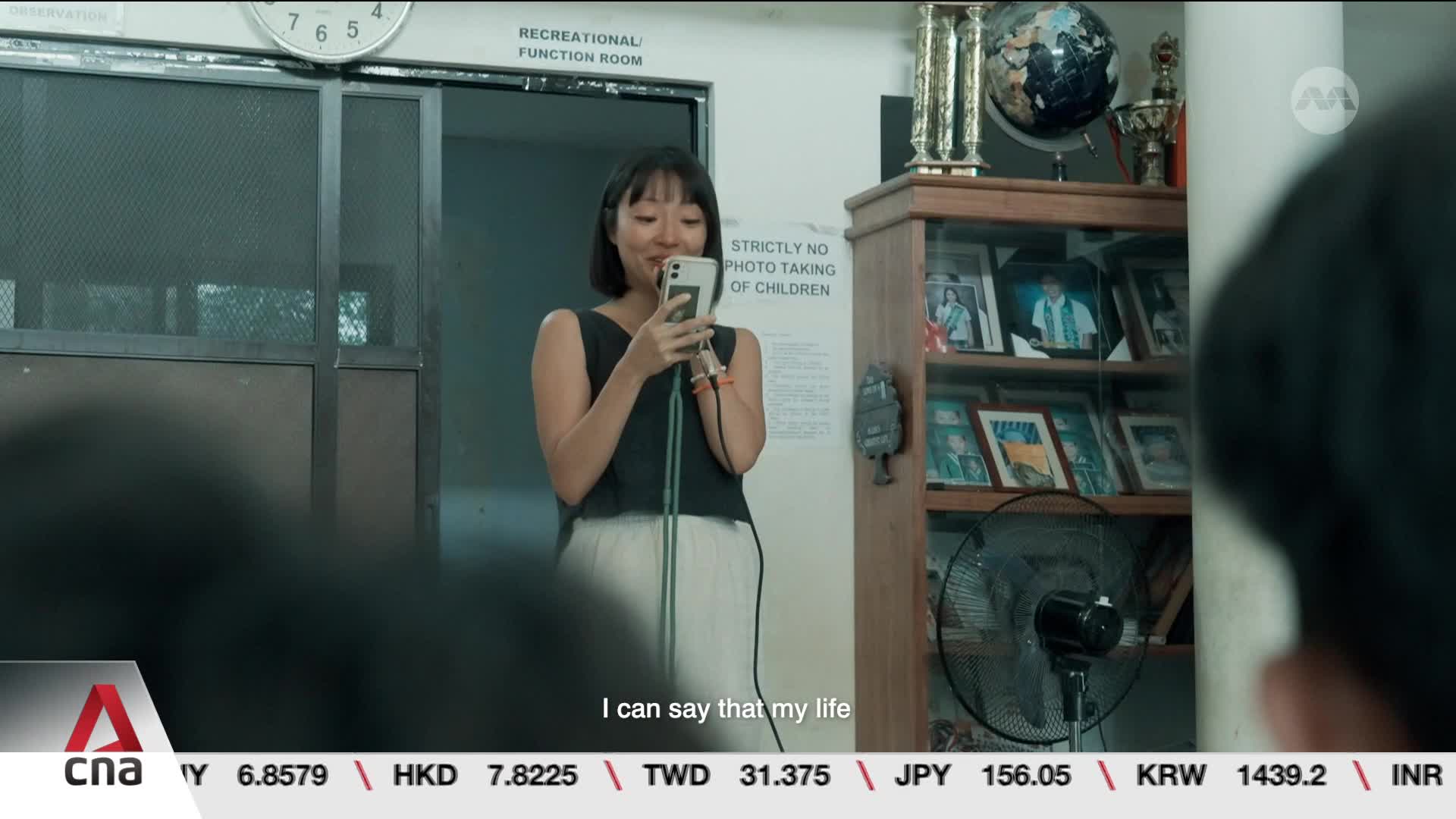 A woman stands holding her phone, recording something. A sign behind her reads "STRICTLY NO PHOTO TAKING OF CHILDREN."