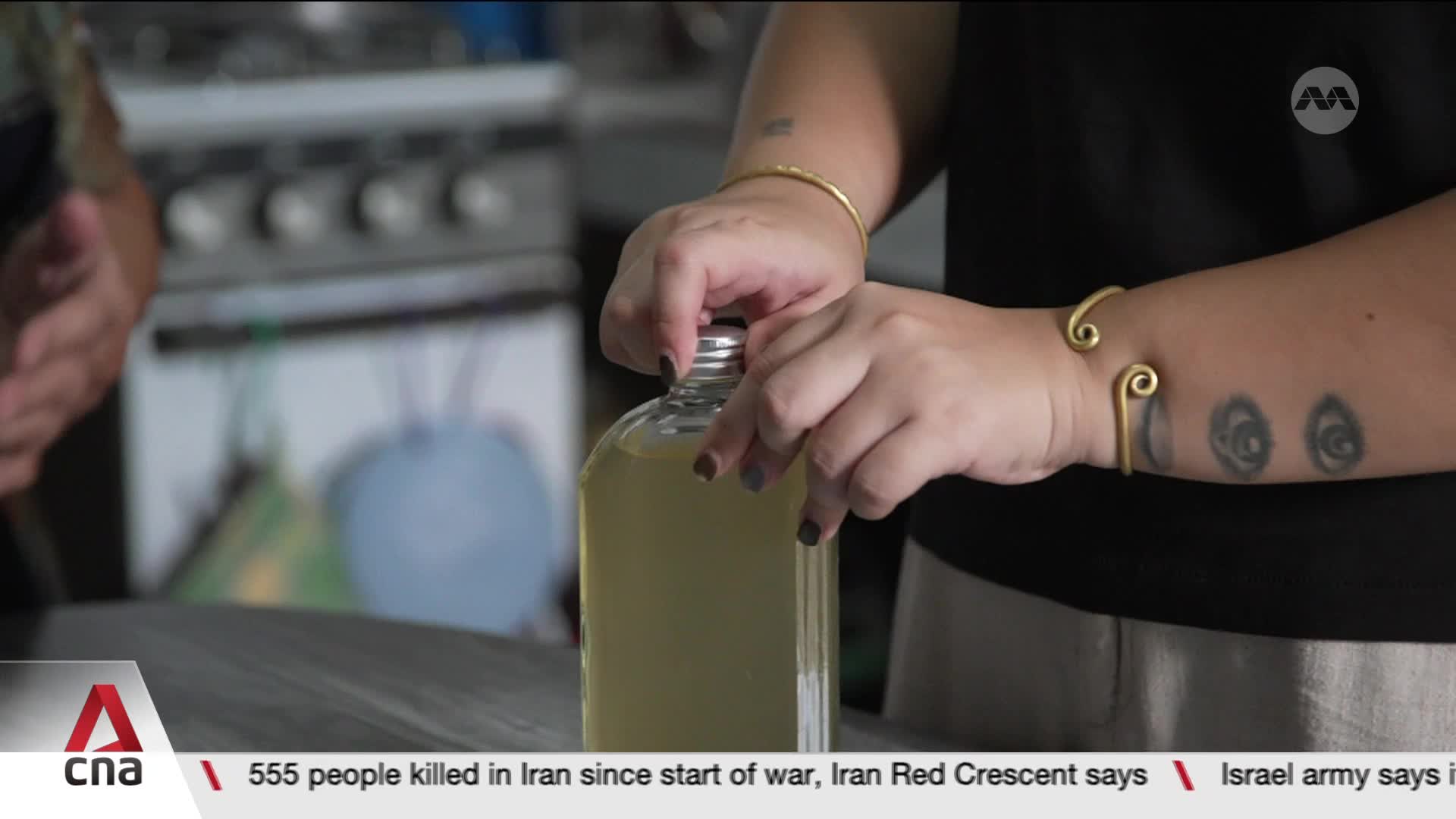 Fingers twist the silver cap of a clear glass bottle filled with pale yellow liquid. The CNA logo appears in the corner, a reminder of the Singaporean broadcast.