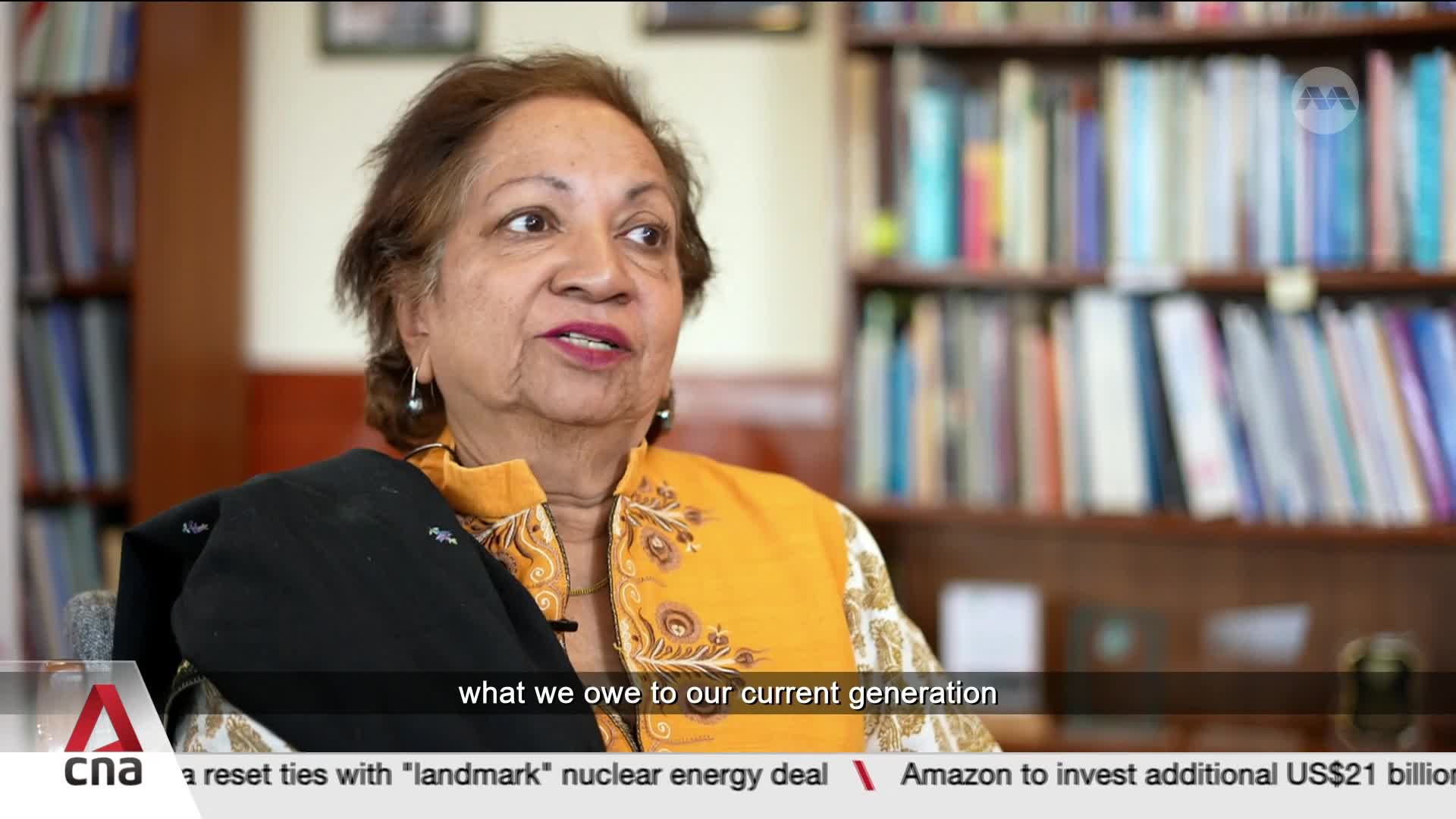 A woman with warm skin and dark hair speaks, her orange and gold patterned top a vibrant contrast to the dark shawl draped over her shoulder. Behind her, bookshelves filled with colorful spines create a backdrop, hinting at a life of learning and reflection.