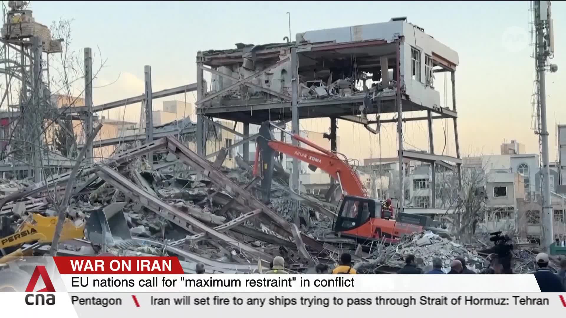 An orange excavator digs through a mountain of rubble. A partially collapsed building looms behind it.