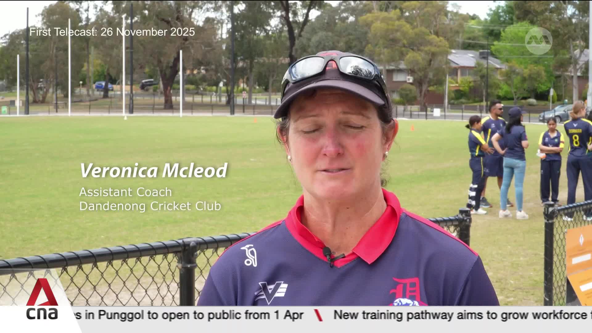 A group of cricketers in navy and yellow uniforms stand on a grassy field, some chatting while others look towards the camera. Veronica McLeod, Assistant Coach for the Dandenong Cricket Club, speaks directly to the viewer.