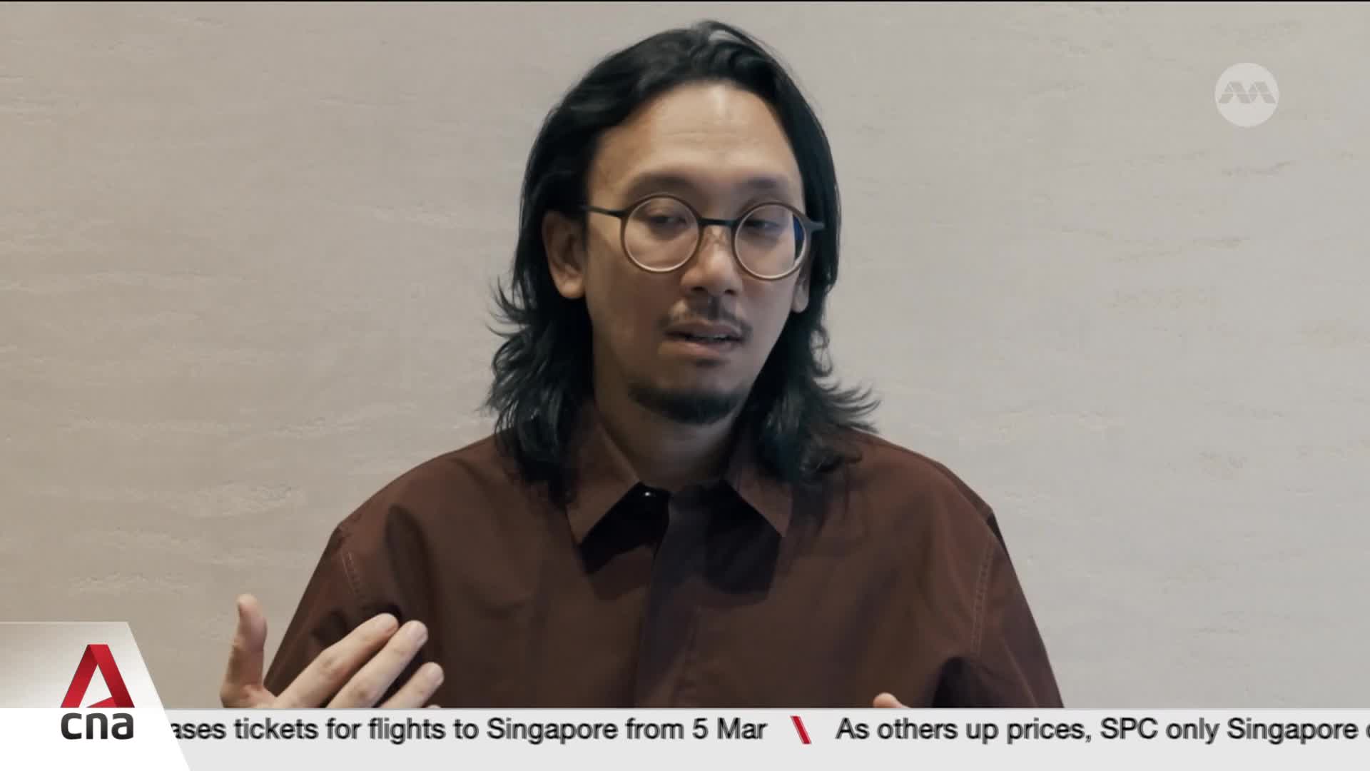 A man with long, dark hair and round glasses speaks, gesturing with his hands. A ticker at the bottom mentions Singapore and CNA.