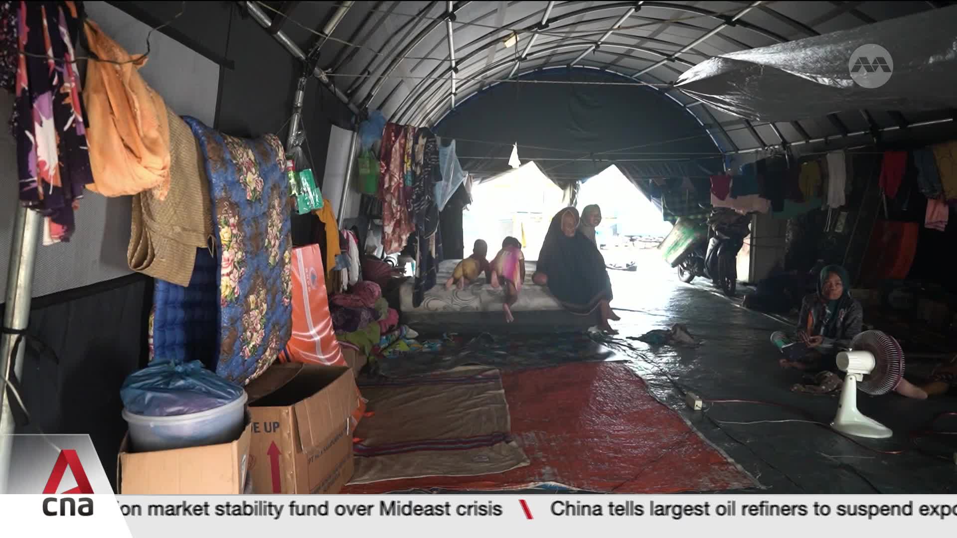 A family sits on a makeshift bed inside a large tent, surrounded by personal belongings. Clothes hang from lines strung across the dark interior, and a small fan whirs near a seated woman.