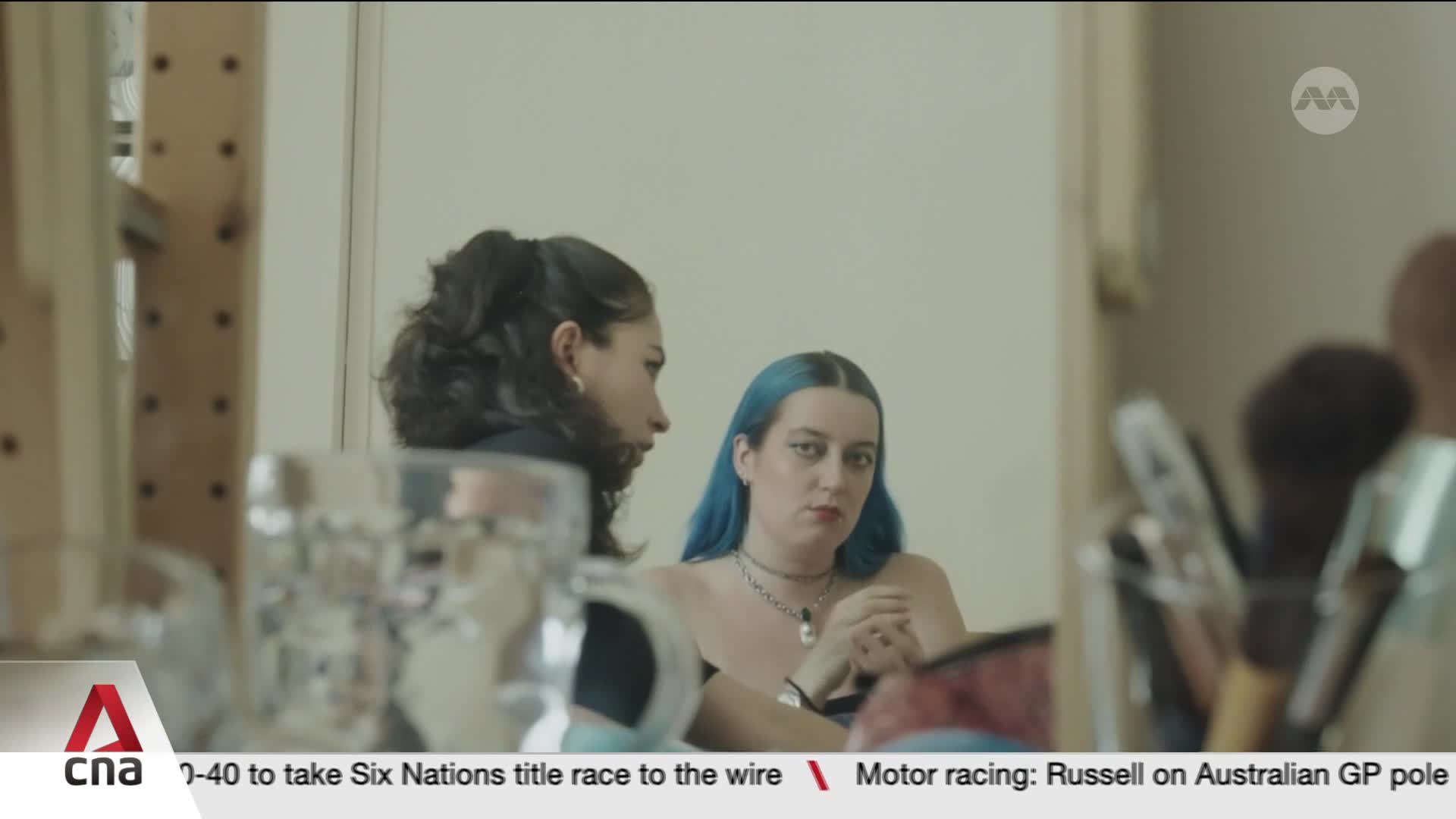 Two young women sit at a vanity, one with dark hair pulled back, the other with bright blue hair. A collection of makeup brushes stands in a container to the right, reflecting the soft light.