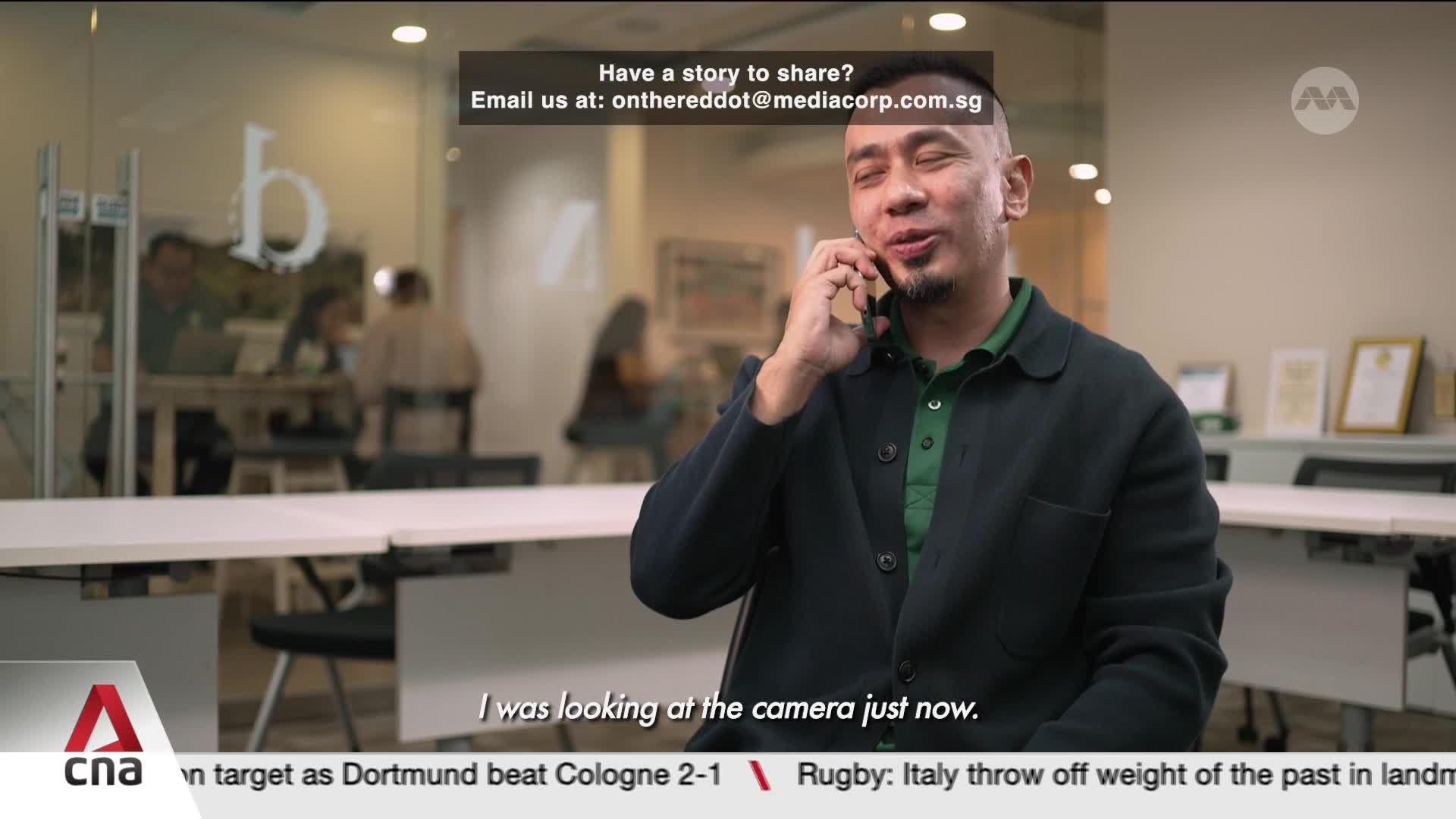 A man in a dark jacket and green collared shirt speaks on his phone. Behind him, through a glass partition, people work at desks in a modern office setting.