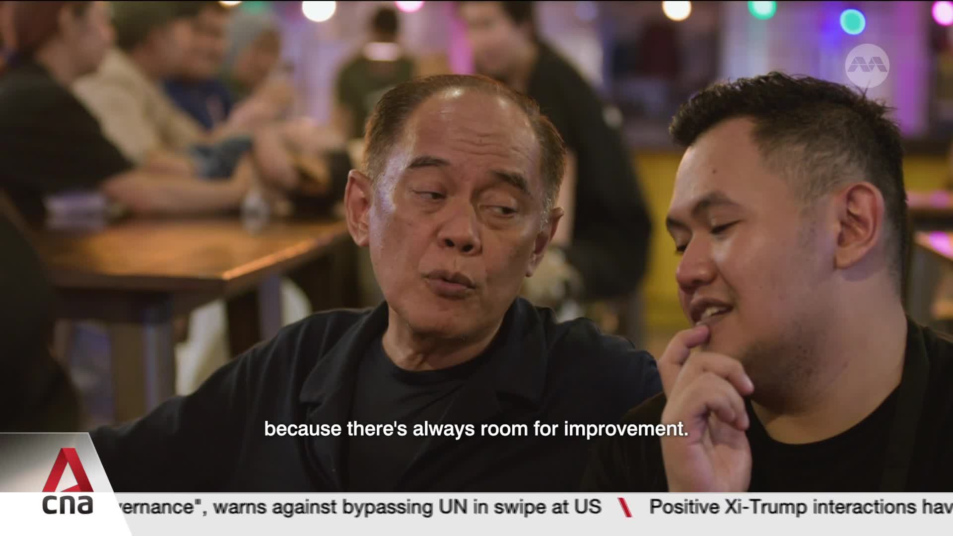 An older man with a dark shirt leans in, speaking intently to a younger man beside him. Behind them, blurred figures and ambient lights suggest a lively setting, perhaps a restaurant in Singapore.