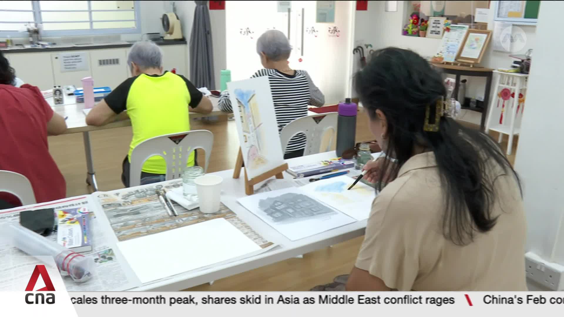 A woman with dark hair pulled back in a ponytail carefully paints on a piece of paper, her brush poised over the surface. Across the table, others are also engaged in their artwork, their focus evident in their posture.