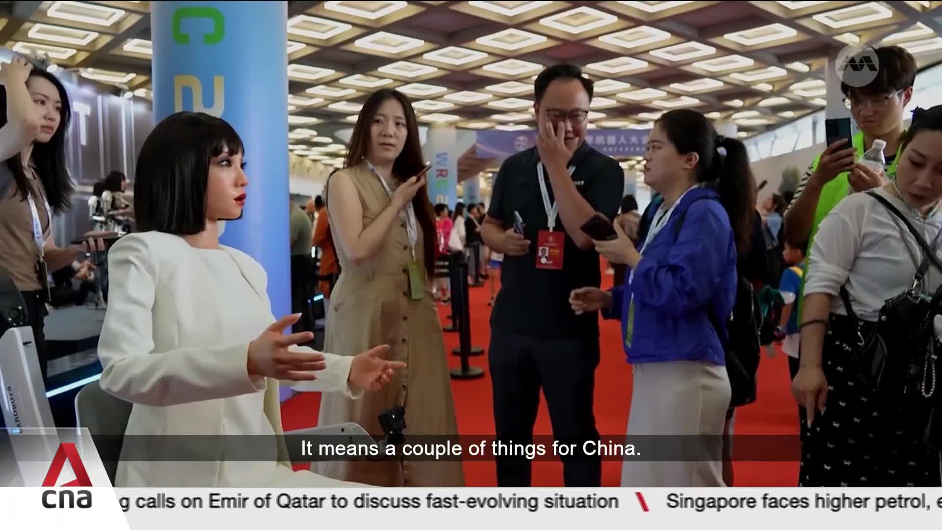 A lifelike robot in a white suit sits attentively, its hands gesturing slightly. Nearby, a group of people are engaged in conversation, some holding phones.