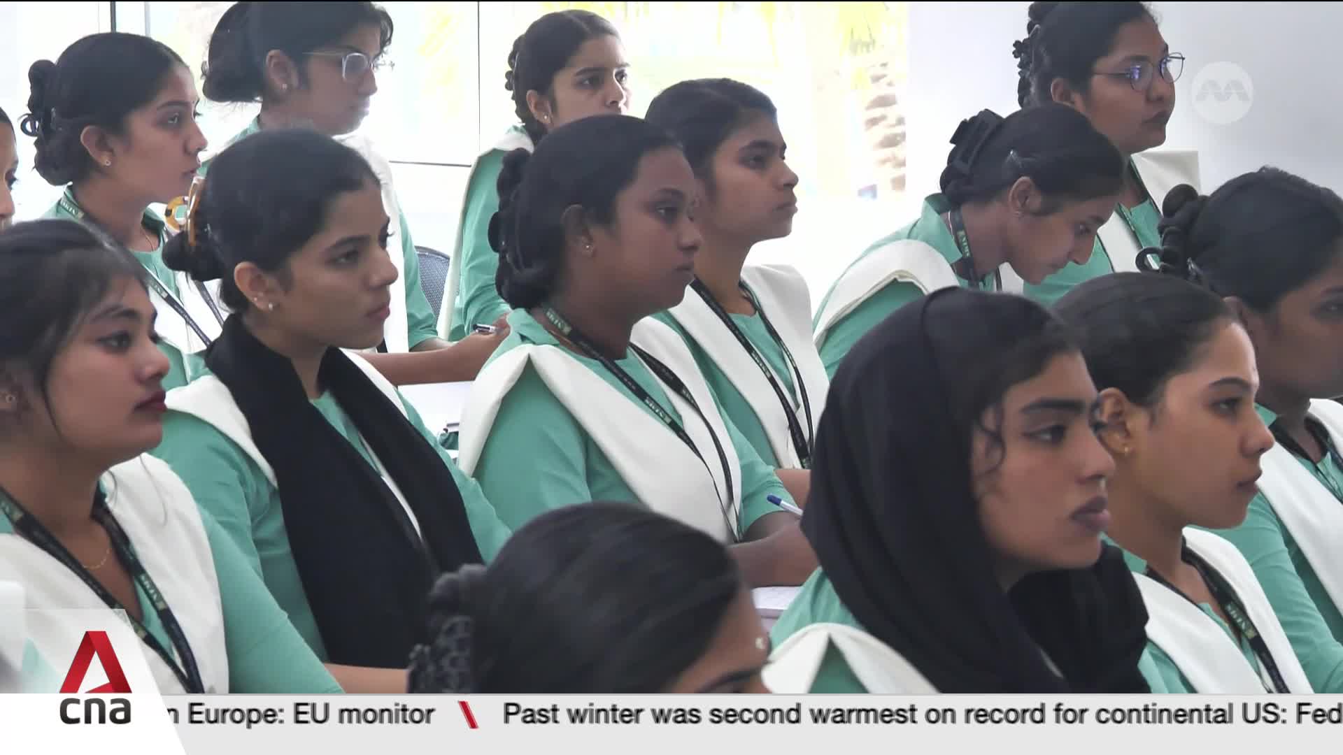 A group of young women, dressed in matching teal and white uniforms, sit attentively. Their gazes are fixed forward, as if listening to a speaker or watching a presentation unfold.