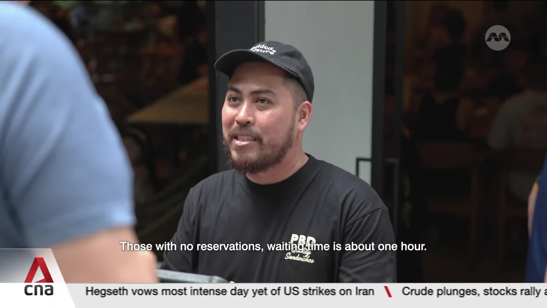 A man in a black t-shirt and cap speaks, his beard visible. Behind him, the interior of a busy establishment in Singapore is dimly lit.