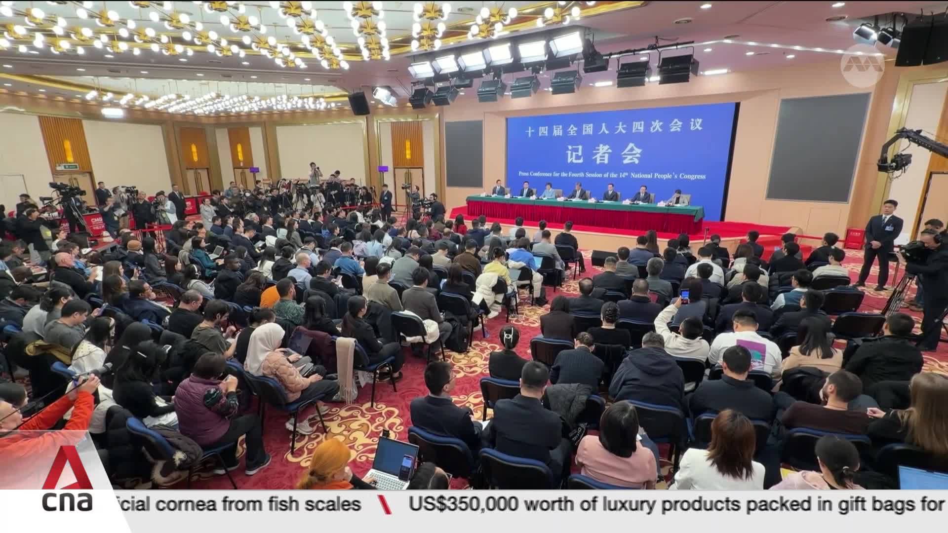 A large crowd of reporters fills this hall, their attention fixed on the panel seated at the front. Cameras and microphones are positioned to capture every word from the press conference.