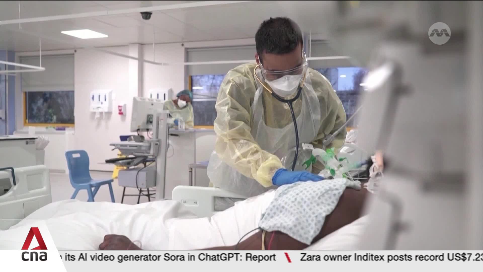 A healthcare worker in full protective gear leans over a patient's bed, stethoscope in hand. Another masked individual in similar attire works at a medical station in the background.