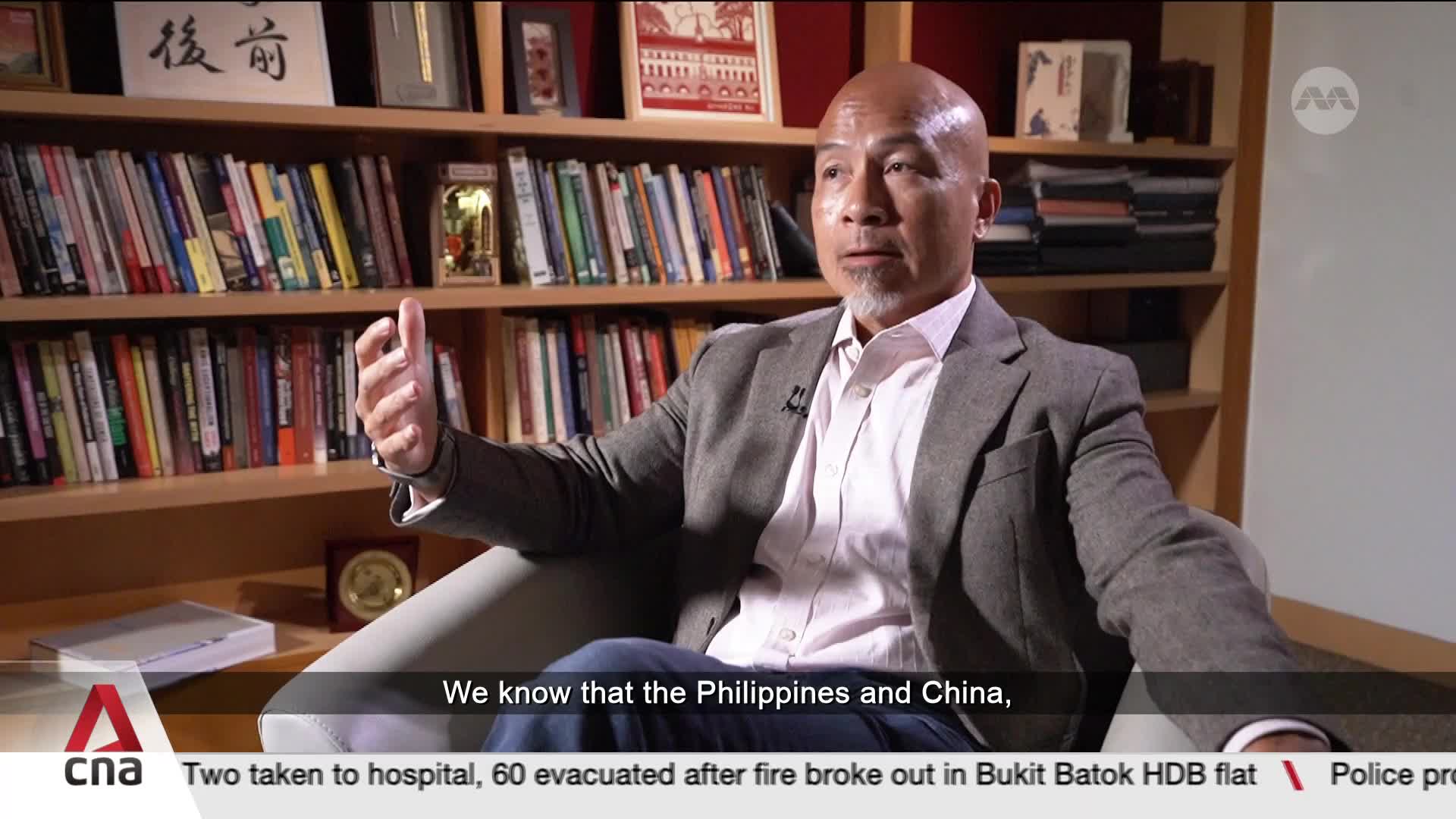 A man in a grey jacket gestures with his hands while speaking, his gaze directed slightly to his left. Behind him, bookshelves are filled with rows of books, with a CNA chyron at the bottom of the screen.