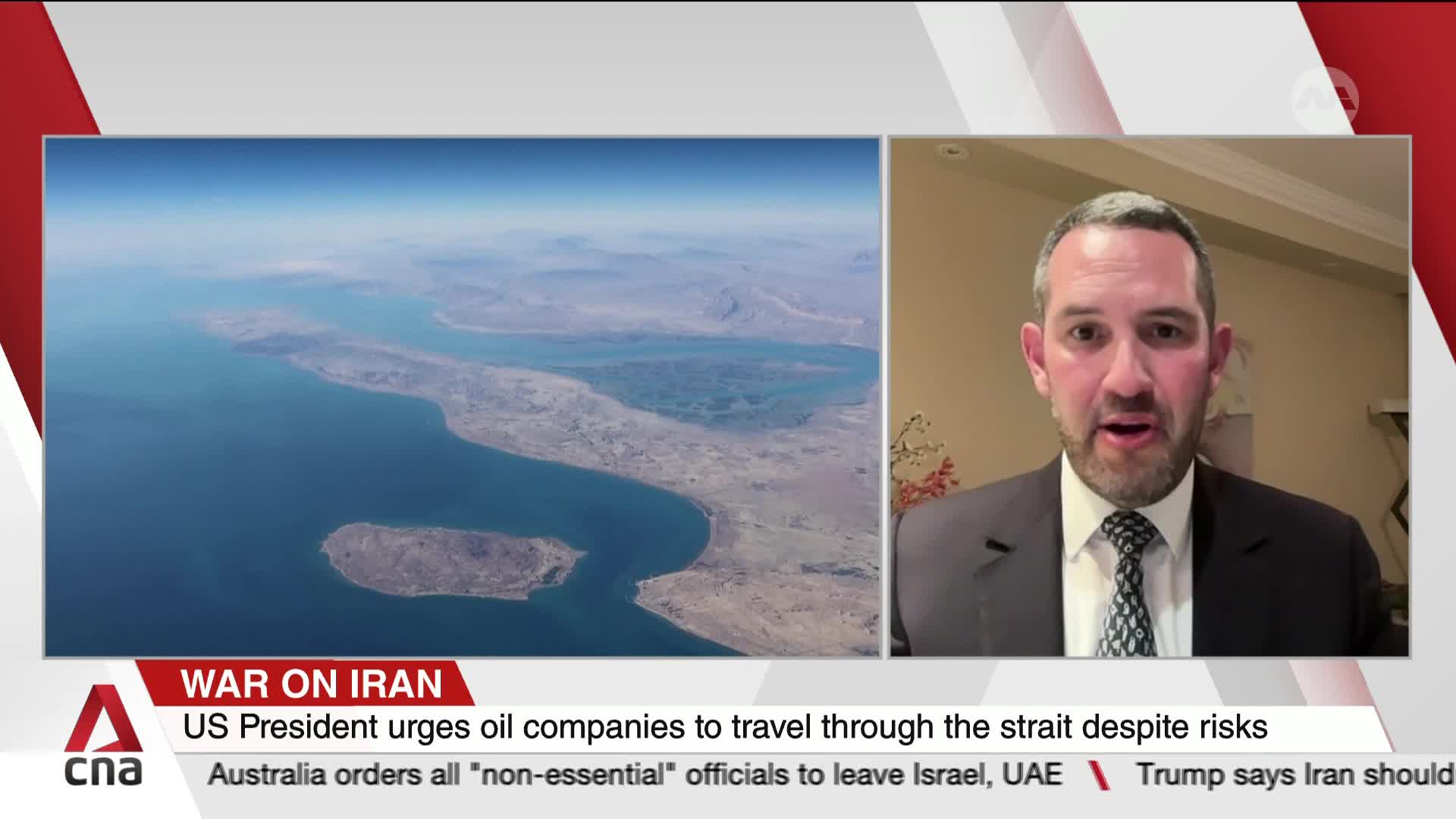 A man in a suit speaks directly to the camera, his face framed by a warm, indoor setting. Beside him, an aerial view shows a vast expanse of blue water meeting arid, mountainous land.