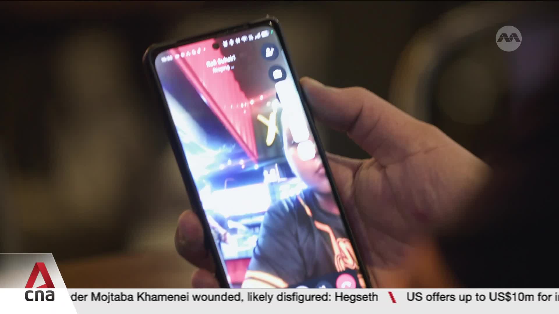 A hand holds a phone displaying a video call. On the screen, a person smiles back, their background a blur of Singaporean neon lights.