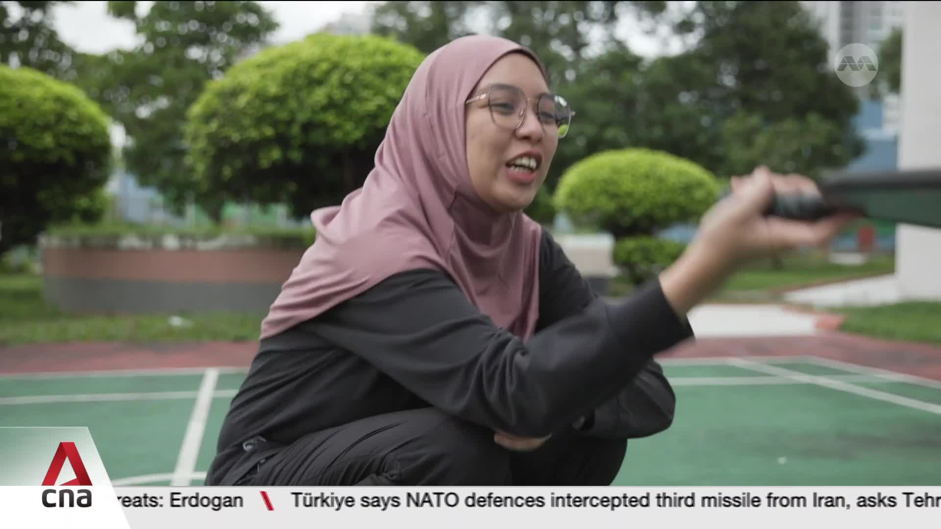 A woman in a pink hijab and glasses crouches on a green court, swinging a badminton racket. She's mid-swing, her mouth open as if speaking.