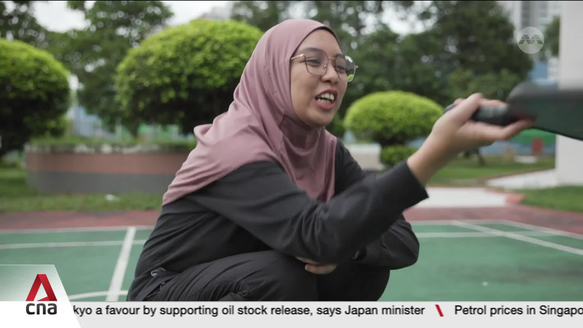 A young woman in a pink hijab and black athletic wear crouches on a green court, holding a dark paddle. She's mid-sentence, her mouth open as she gestures with the paddle.