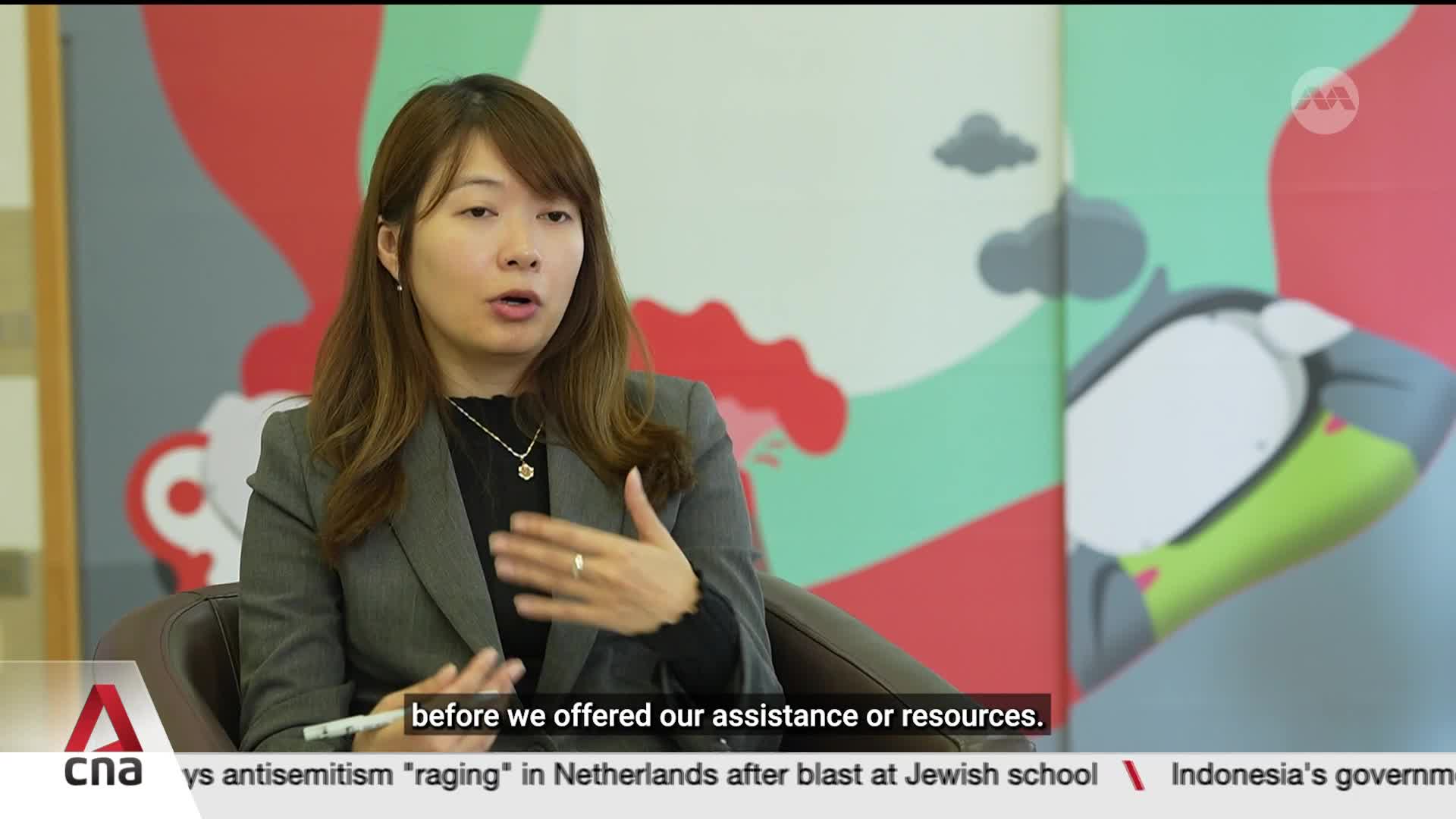 A woman in a grey blazer speaks, gesturing with her hands. Behind her, a colorful mural features abstract shapes and stylized clouds.