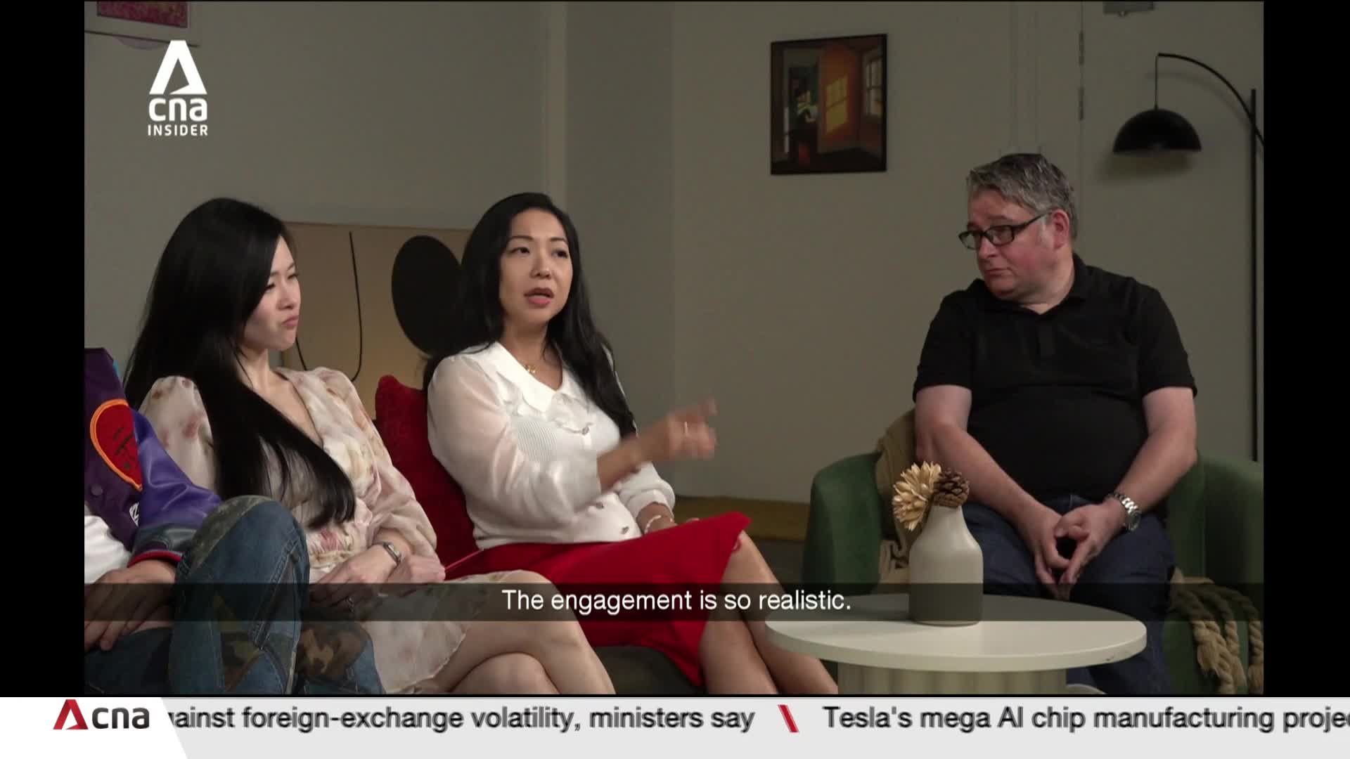 A woman in a white blouse and red skirt gestures animatedly, speaking to a man in a black polo shirt. Another woman sits beside her, listening intently. A woman in a white blouse and red skirt gestures animatedly, speaking to a man in a black polo shirt. Another woman sits beside her, listening intently.