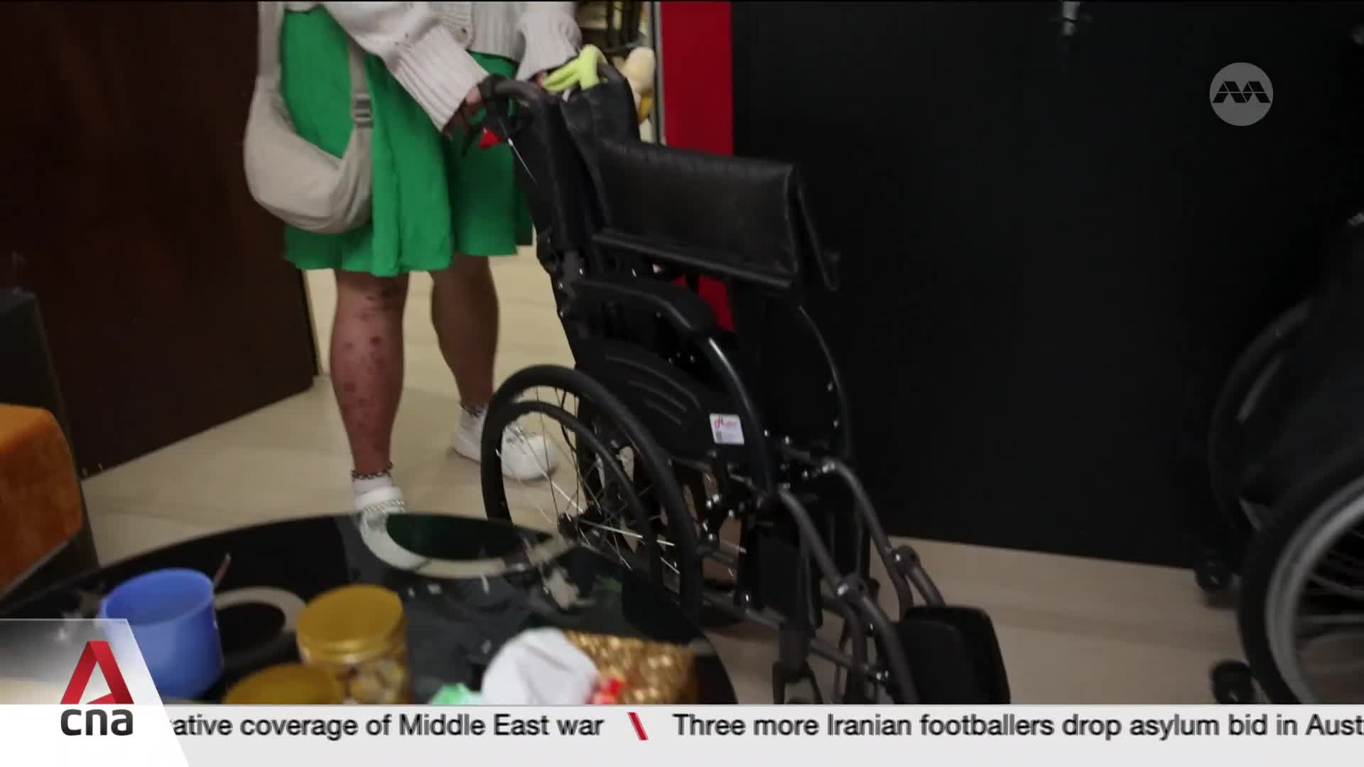 A person in a green dress pushes a wheelchair. The scene is indoors, with a dark background and a tiled floor. A person in a green dress pushes a wheelchair. The scene is indoors, with a dark background and a tiled floor.