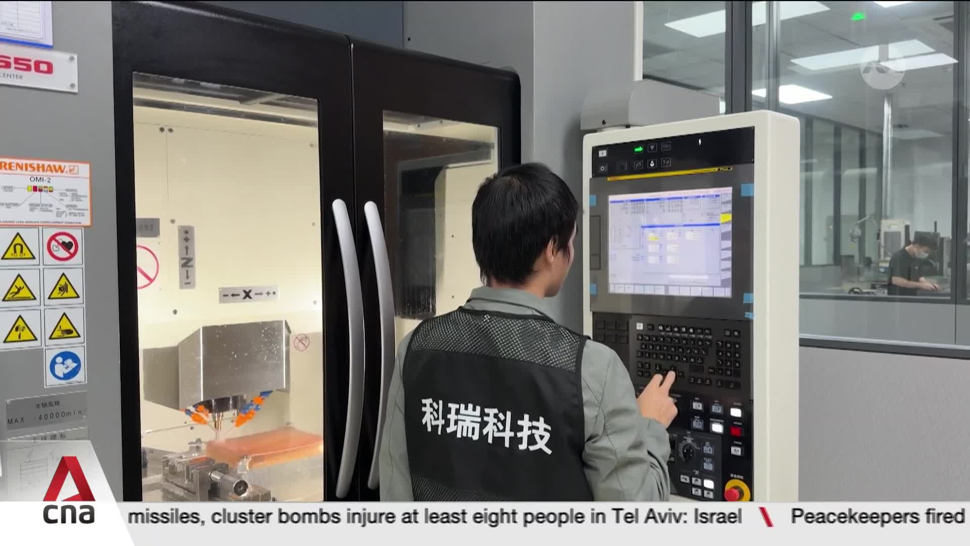 A technician in a vest with Chinese characters operates a complex control panel. Inside the machine, a bright orange component is being worked on. A technician in a vest with Chinese characters operates a complex control panel. Inside the machine, a bright orange component is being worked on.