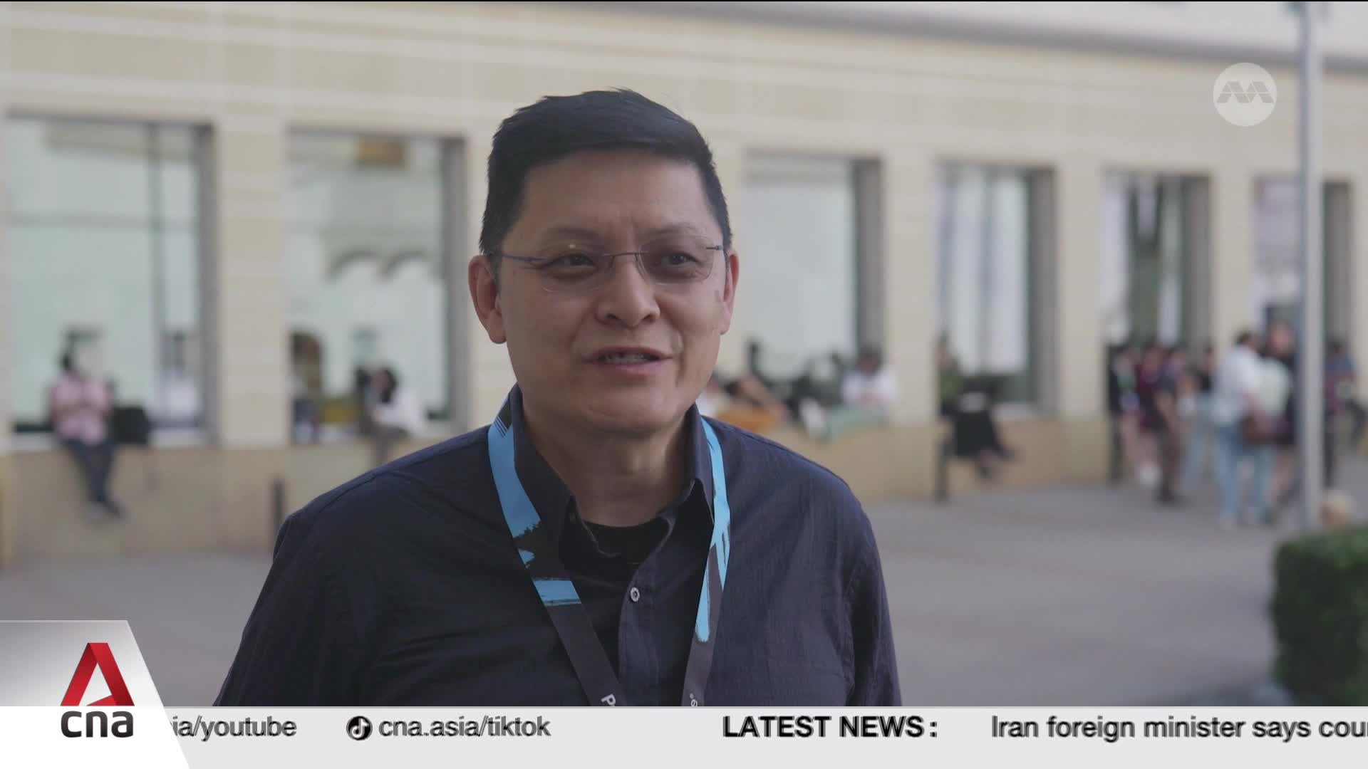 A man in a dark shirt with a lanyard speaks directly to the camera. Behind him, a crowd of people mills about in front of a modern building. A man in a dark shirt with a lanyard speaks directly to the camera. Behind him, a crowd of people mills about in front of a modern building.