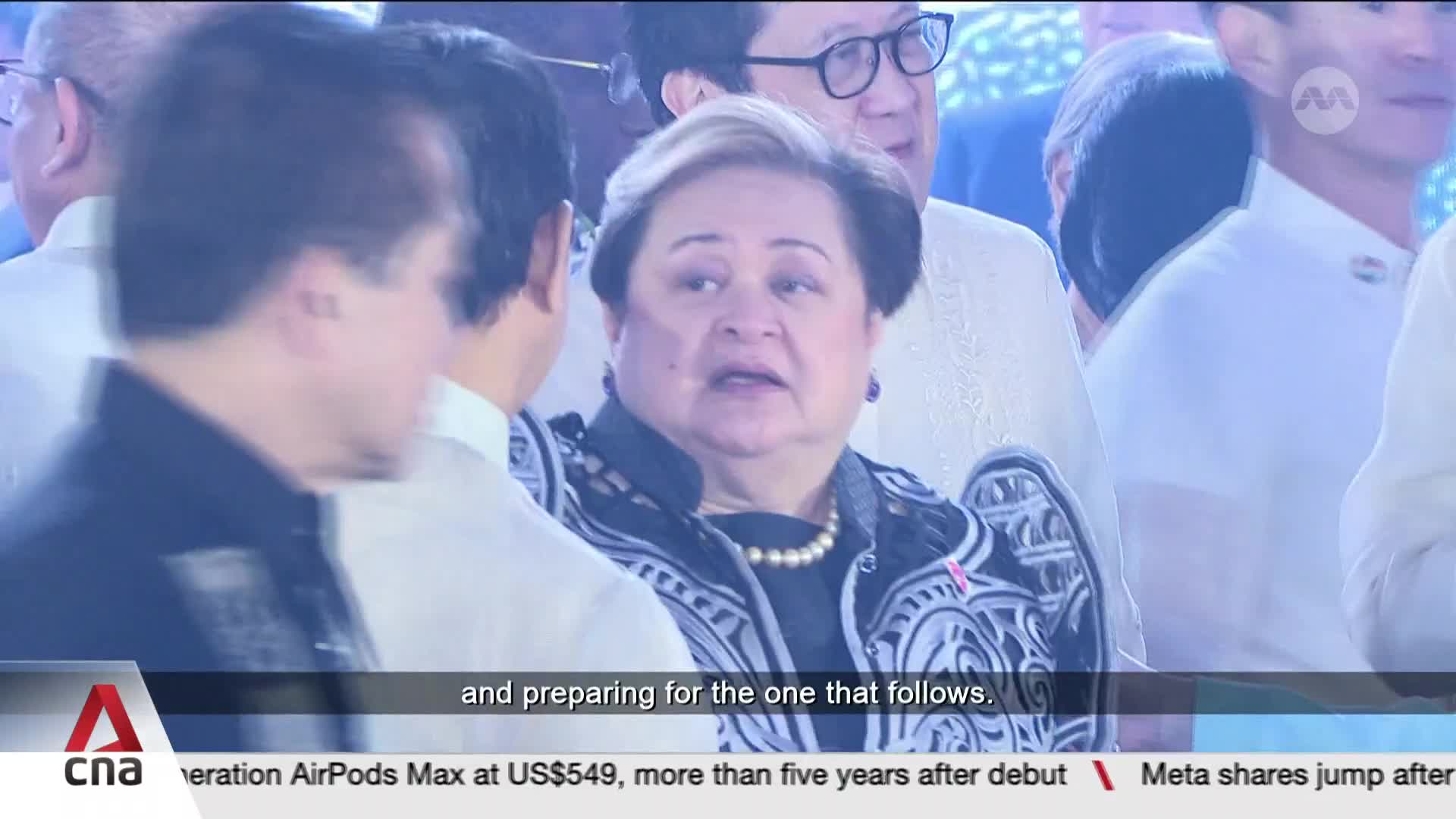 A woman in a black and white patterned jacket, wearing a pearl necklace, speaks with someone to her left. The CNA International logo is visible in the lower corner. A woman in a black and white patterned jacket, wearing a pearl necklace, speaks with someone to her left. The CNA International logo is visible in the lower corner.