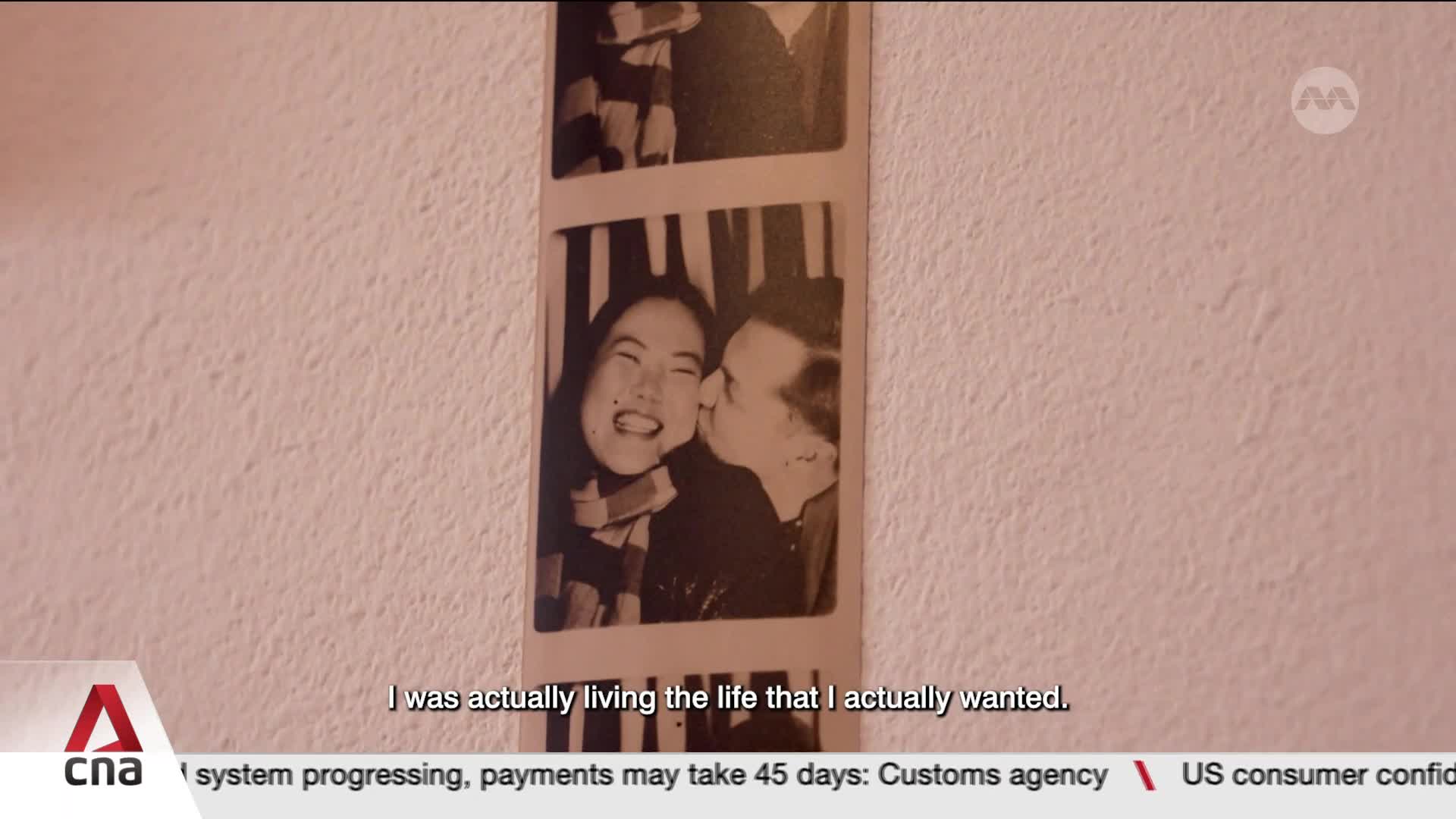 A man gently kisses a woman's cheek in a photo strip, her eyes closed in a joyful expression. The black and white image hangs on a textured wall, a moment captured from Singapore.
