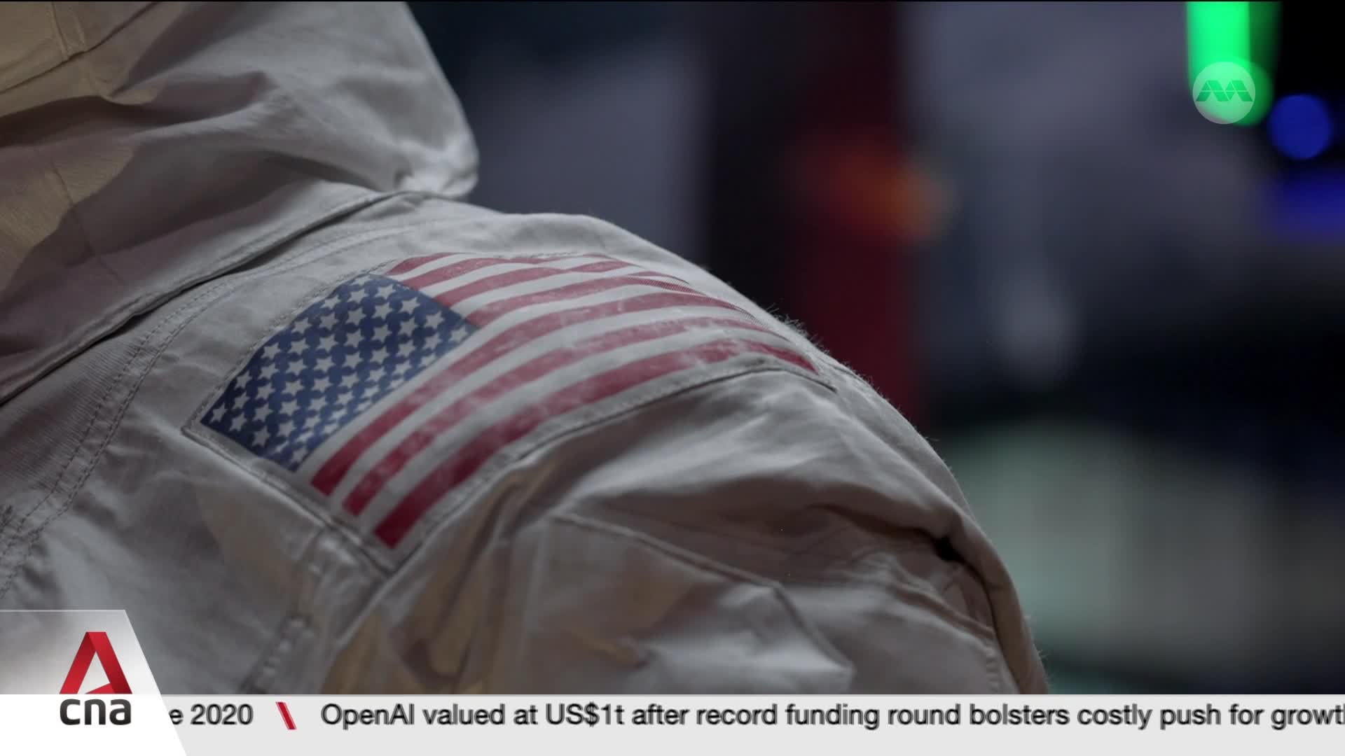 An astronaut's shoulder patch, the American flag, is visible on their suit. The background shows blurred lights, hinting at a Singaporean broadcast from CNA International.