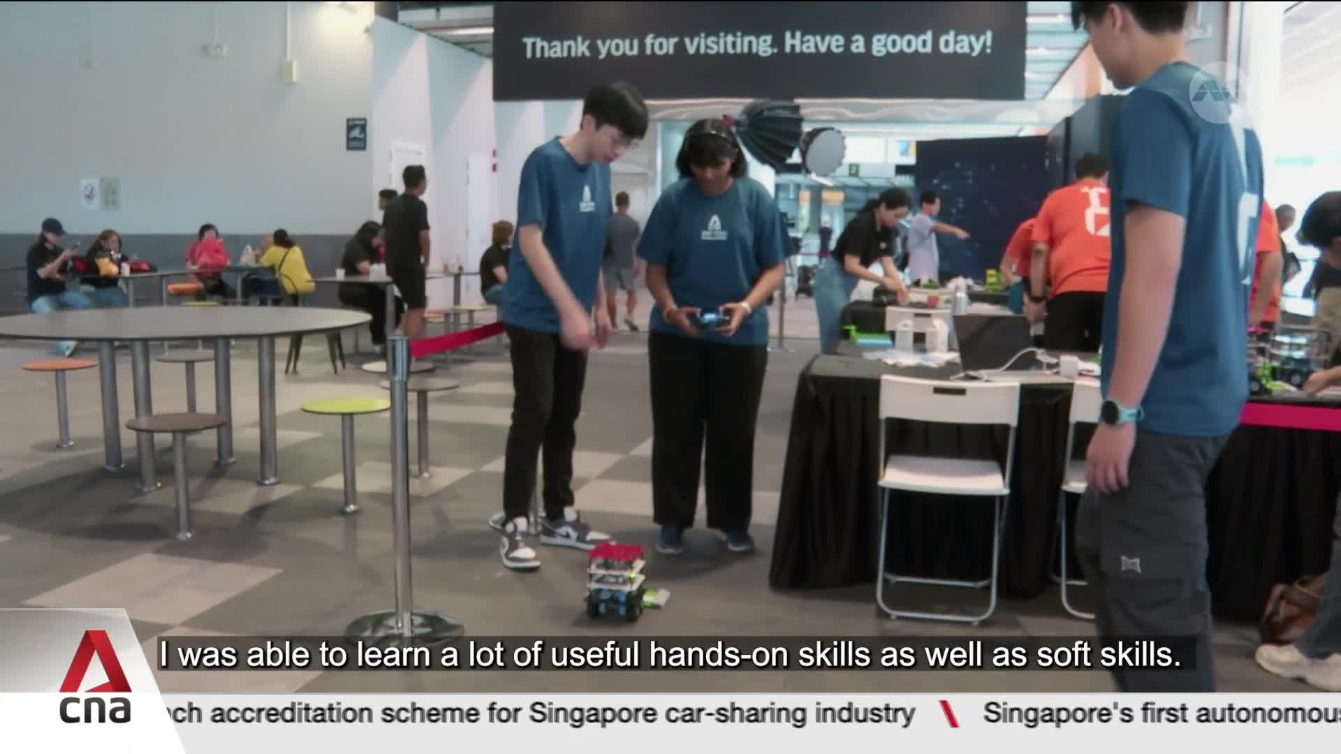 A young man in a blue t-shirt controls a small, multi-colored robot with a remote. Beside him, a woman in a similar shirt watches the robot as it moves across the floor.