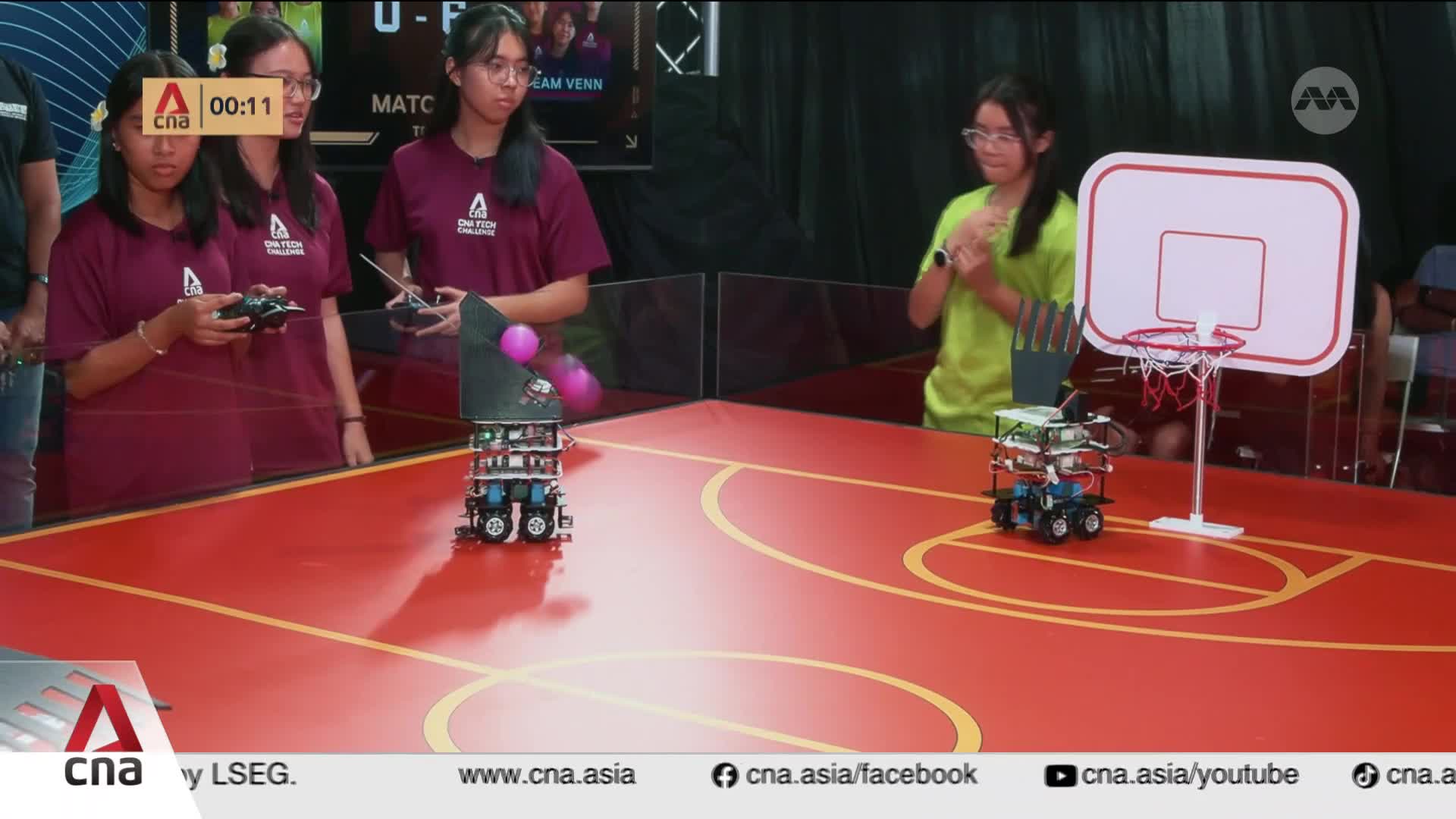 Two robots, controlled by students, navigate a miniature basketball court in Singapore. One robot is positioned near a hoop, while the other, with pink balls atop its structure, moves across the red surface.