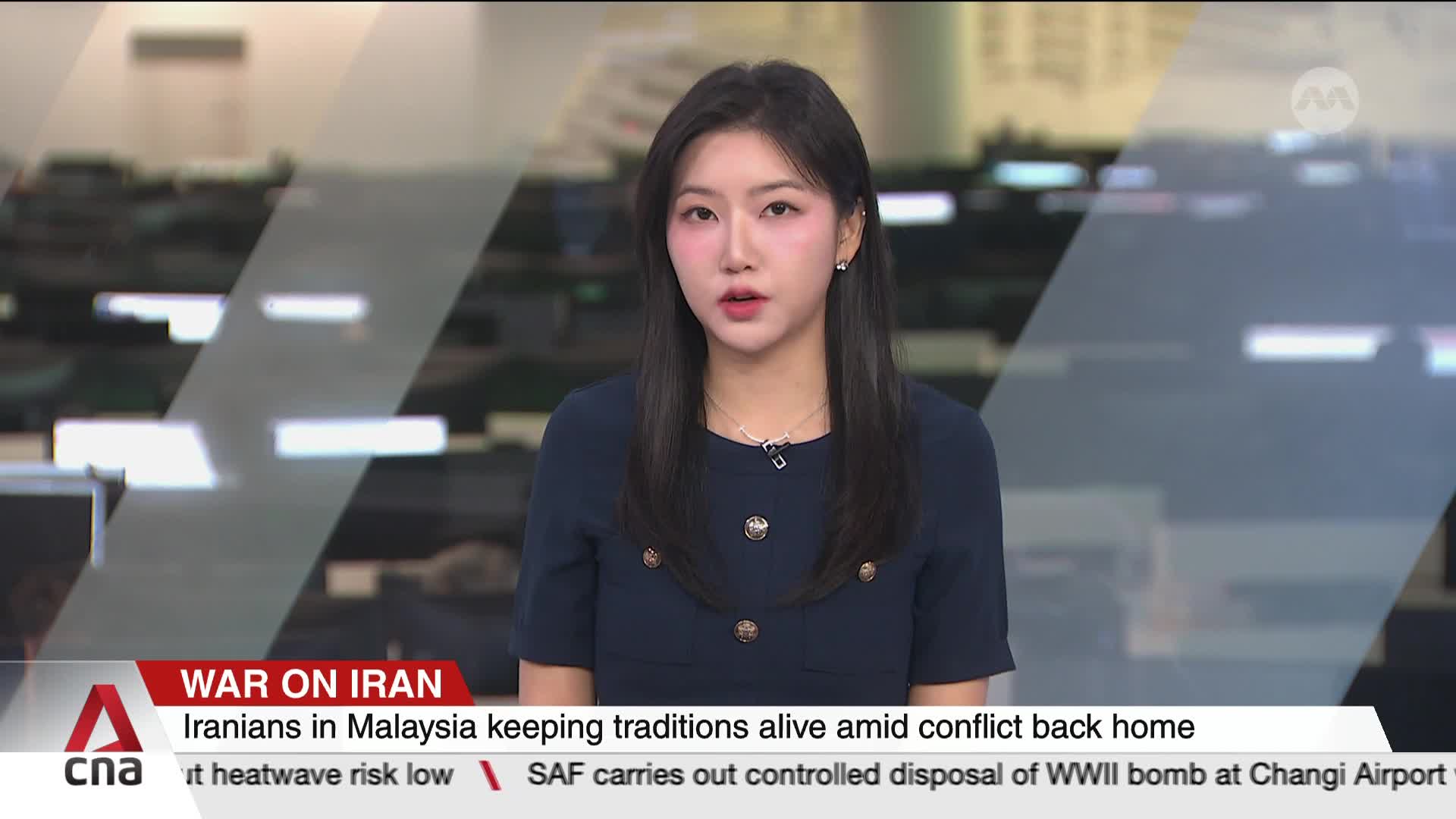 A woman in a dark blue dress stands before a screen showing news headlines. The bottom of the screen displays "WAR ON IRAN" and "Iranians in Malaysia keeping traditions alive amid conflict back home."