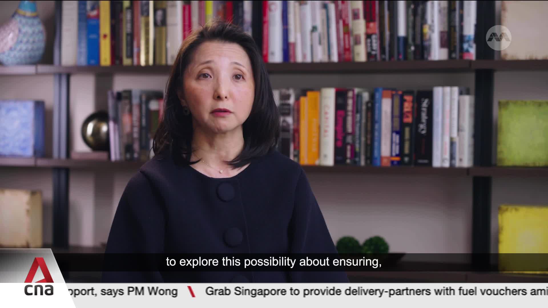 A woman with dark hair, wearing a dark jacket, speaks directly to the camera. Behind her, bookshelves are filled with various titles. A chyron at the bottom displays "CNA" and a news ticker mentions "Grab Singapore."