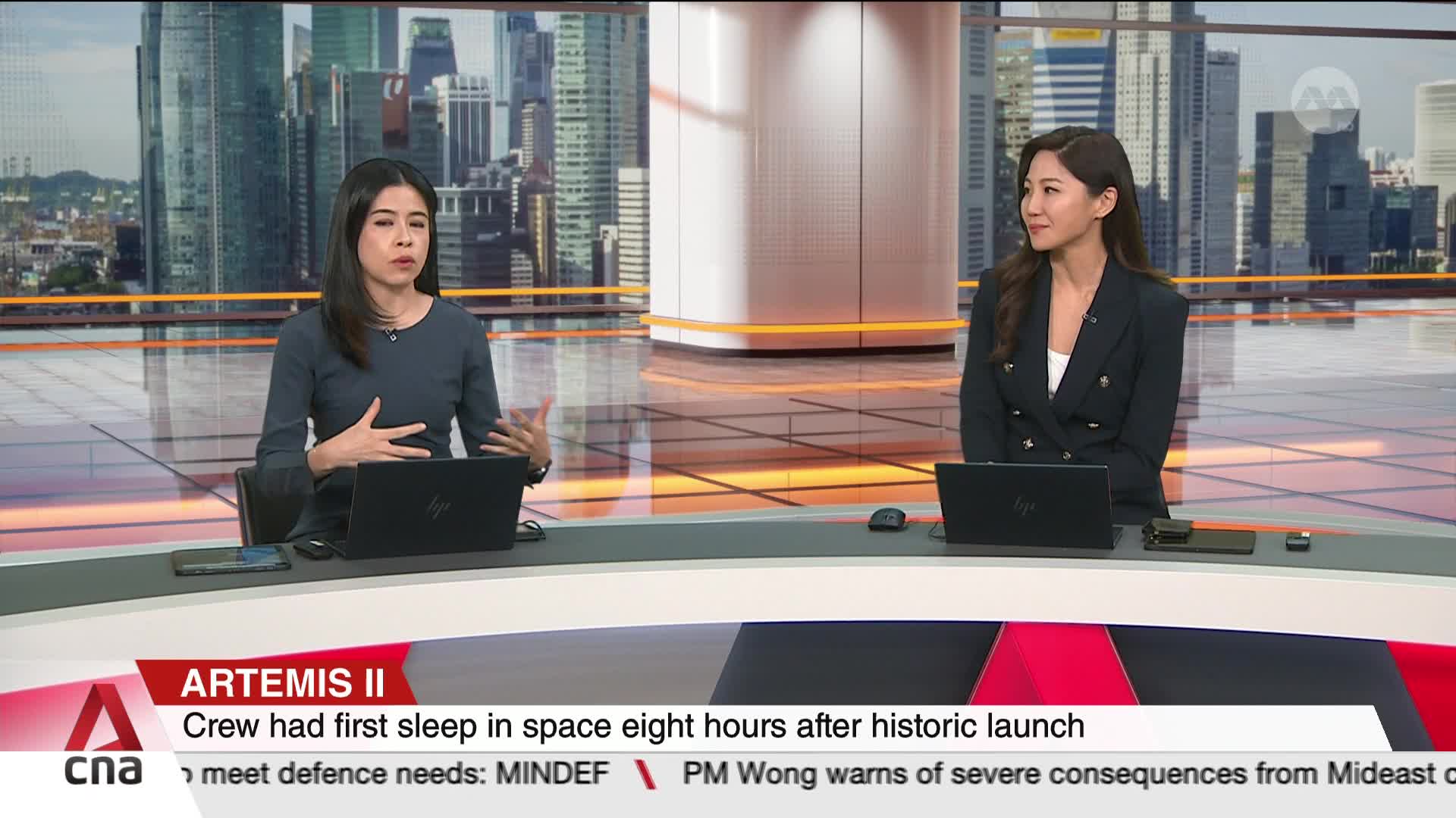 Two women are seated at a desk, speaking to the camera. Behind them, a cityscape of tall buildings is visible, reflecting the bright studio lights.