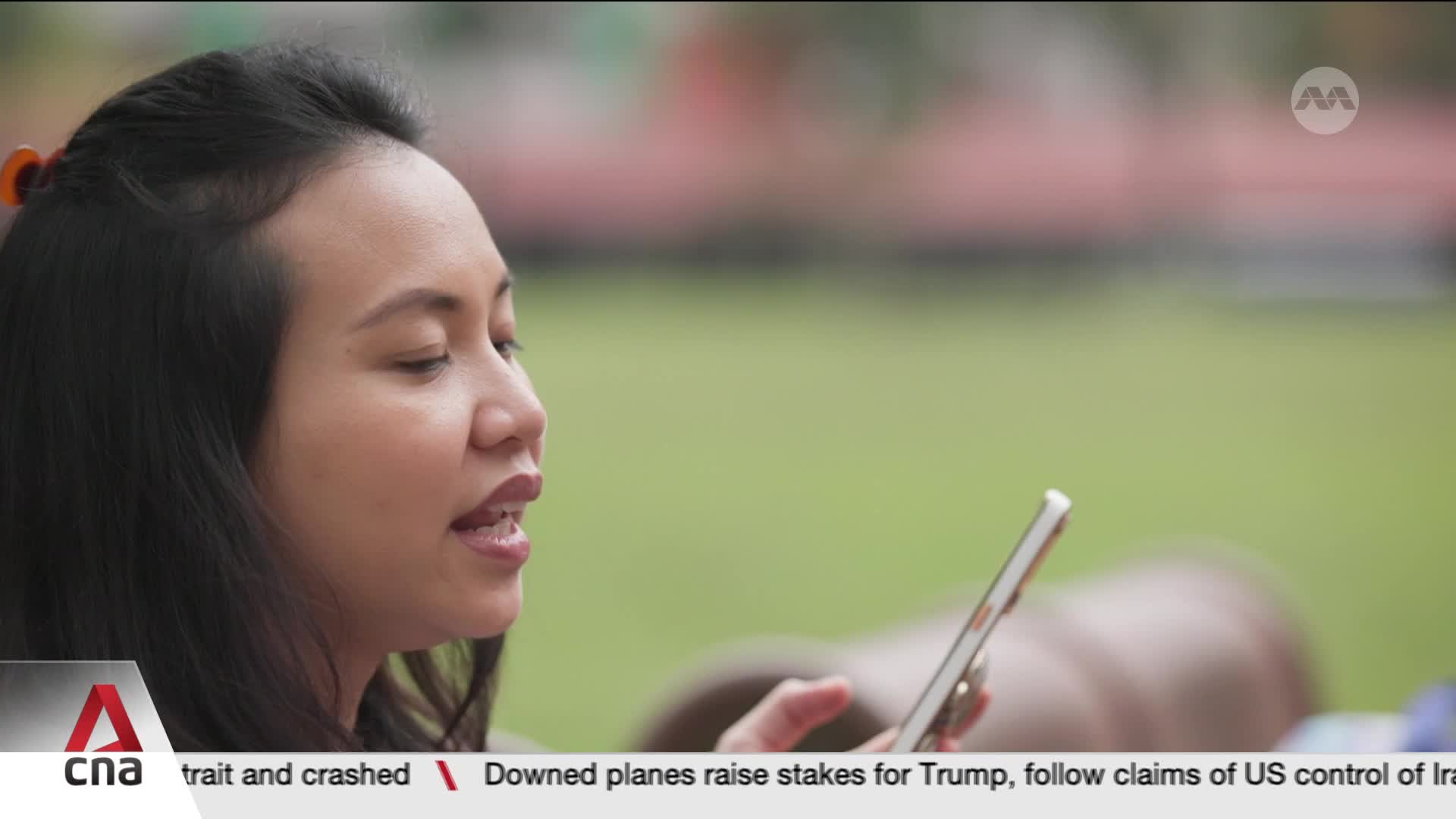 A woman in Singapore is speaking into her phone, her expression engaged. The background suggests an outdoor setting, with a hint of news ticker from CNA International scrolling at the bottom.