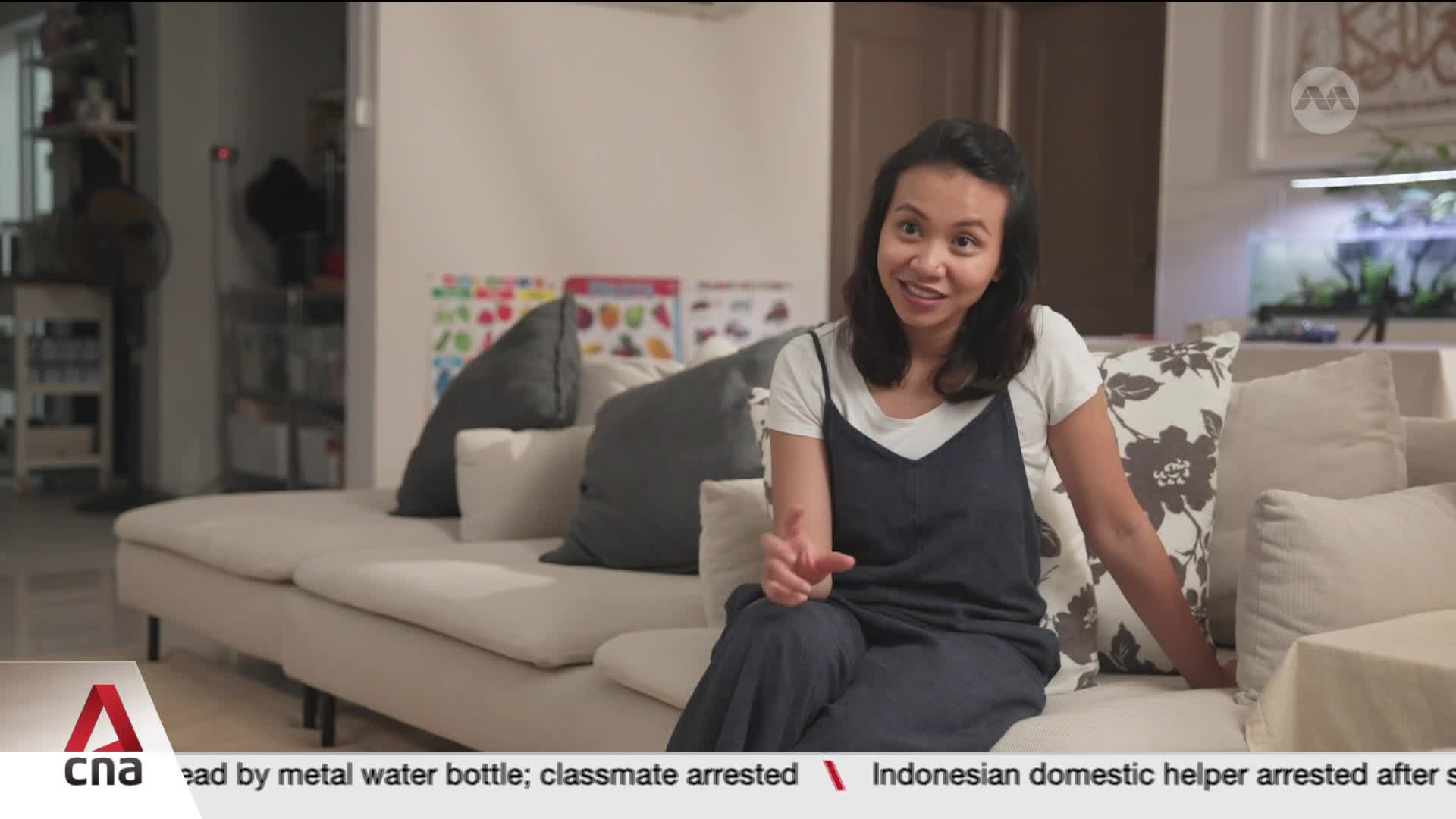 A woman sits on a plush sofa, gesturing as she speaks. Behind her, a brightly colored chart hangs on the wall, and a shelf with various items is visible.