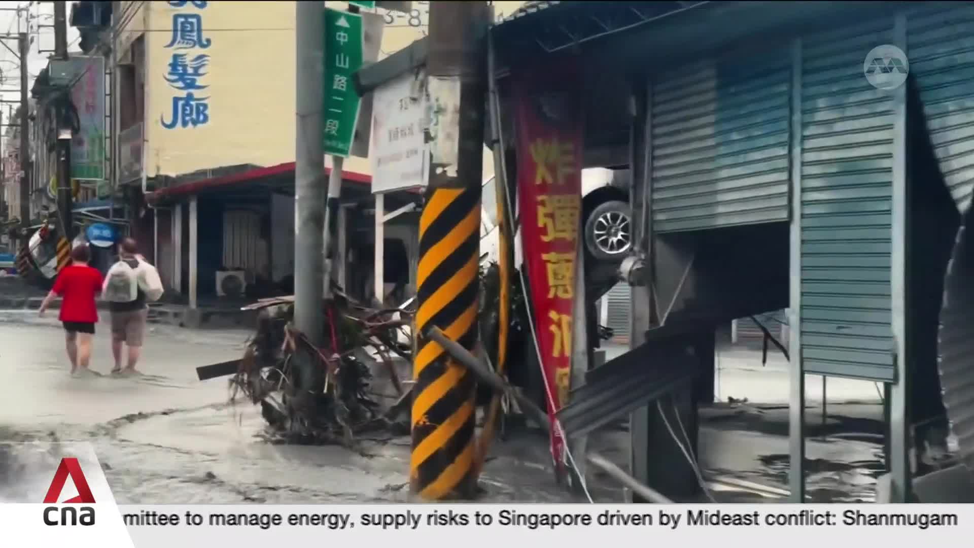A car is wedged precariously in the doorway of a shop, its wheel visible. Two people walk through debris-strewn water on the street, a stark reminder of the recent storm.