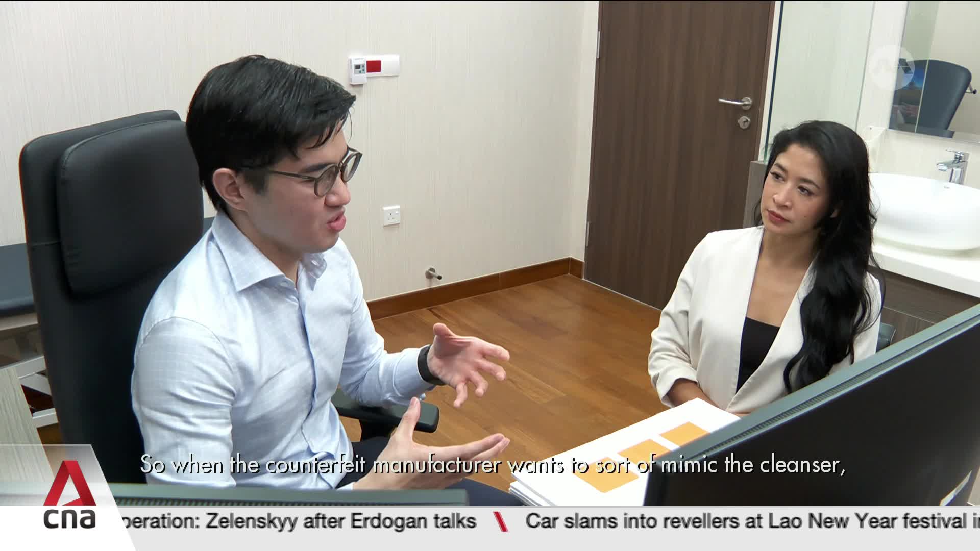 A man in a light blue shirt gestures animatedly while speaking to a woman in a white blazer. They are seated at a desk in a modern office, with a CNA chyron visible at the bottom of the frame.