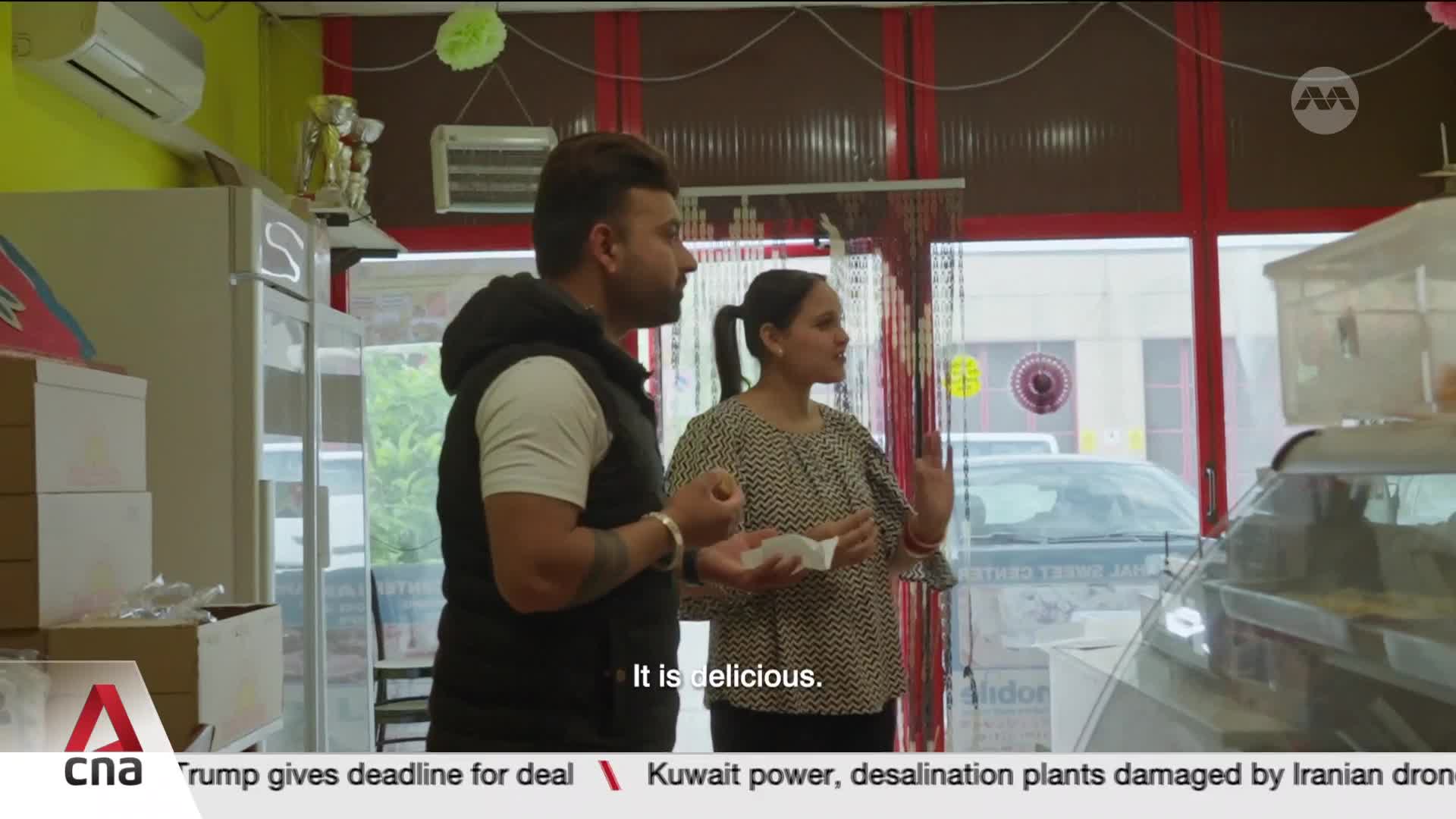 A man and woman stand in a brightly lit shop, holding small white pieces of food. The woman gestures with her free hand as she speaks, and the man listens intently. A man and woman stand in a brightly lit shop, holding small white pieces of food. The woman gestures with her free hand as she speaks, and the man listens intently.