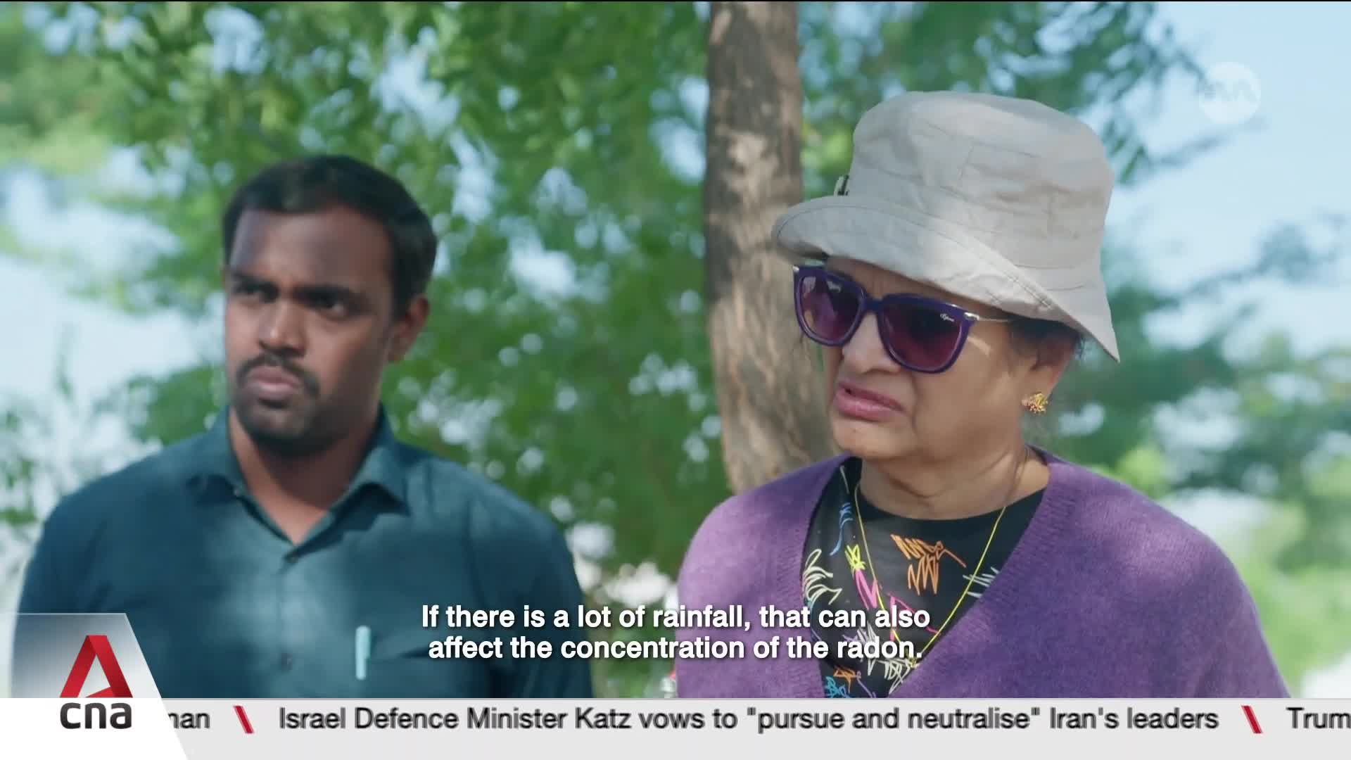 A man in a teal shirt looks intently to his left, while a woman in a purple cardigan and a wide-brimmed hat speaks with a concerned expression. Their conversation is being subtitled by CNA.