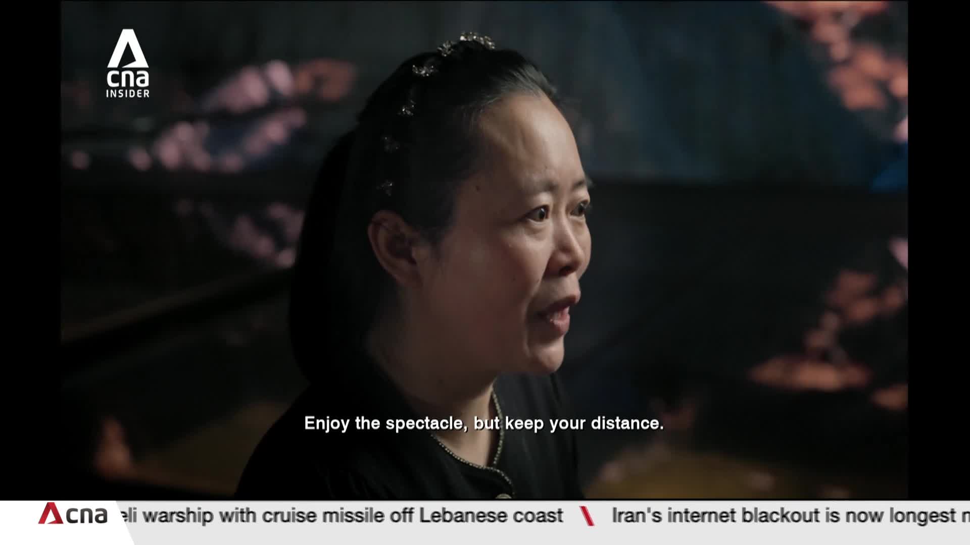 A woman with her hair pulled back speaks, a delicate band adorning her forehead. Behind her, a soft, out-of-focus light display hints at a Singaporean spectacle.