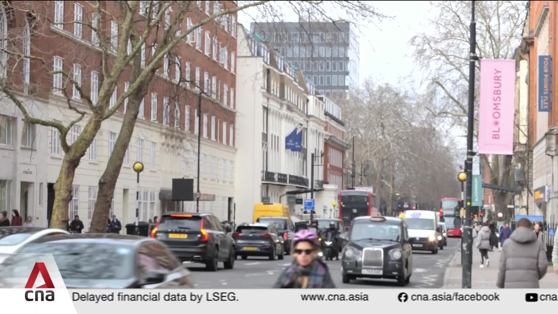 Cars and a motorcycle navigate a busy street lined with buildings. A pink banner advertising "Bloomsbury" hangs from a lamppost.