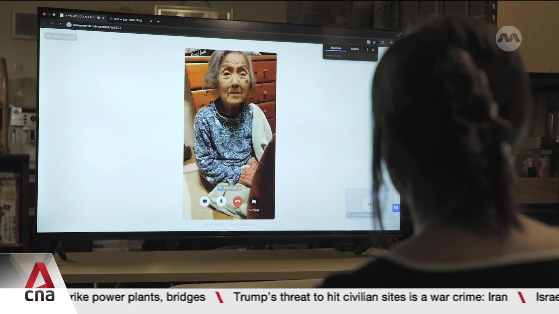 An elderly woman with grey hair, wearing a blue patterned top, smiles from a video call on a large monitor. The screen displays the CNA International logo in the corner, hinting at a broadcast from Singapore.