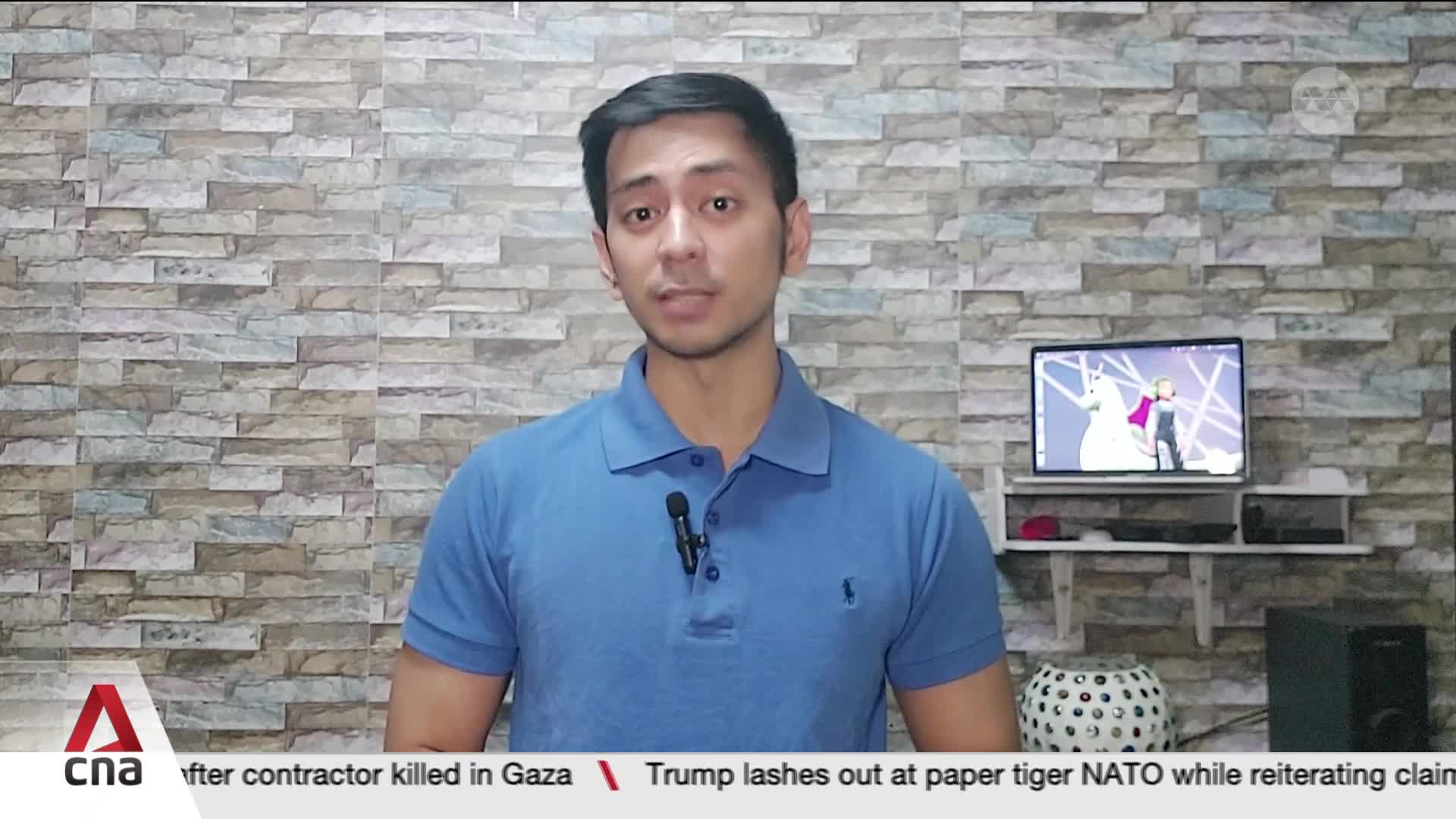 A man in a blue polo shirt stands before a textured wall, speaking into a microphone. On a shelf behind him, a small television displays an animated scene. A news ticker runs along the bottom, showing headlines from CNA.